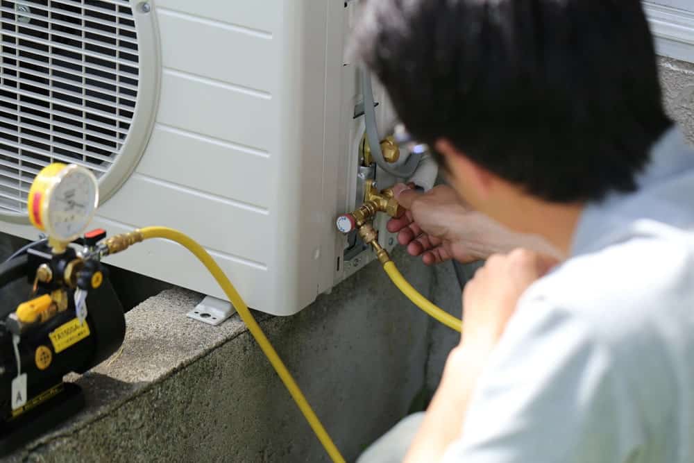 A person checks an air conditioning unit's connections outside, with a gauge and yellow hose attached, suggesting maintenance or repair work.
