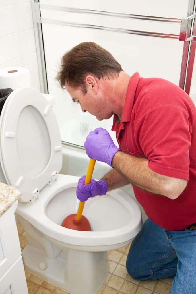 Man in a red shirt and purple gloves uses a plunger on a toilet in a bathroom.