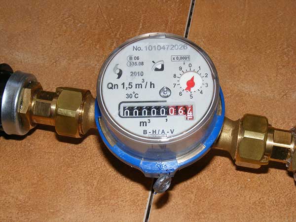 Close-up of a water meter with a reading of 664 cubic meters. The meter is installed on a tiled surface and connected to pipes on either side.