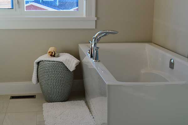 Bathroom scene with a standalone bathtub, a white mat, wicker basket with rolled towel, and a window. The walls are beige and floor tiles are light-colored.