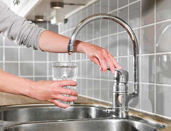 Person filling a glass with water from a kitchen faucet over a sink.