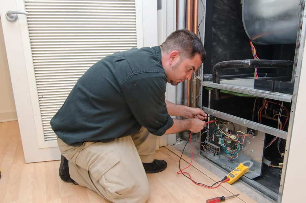 A technician kneels while repairing an HVAC unit, holding tools. Wires and a multimeter are visible, and the setting is indoors on a wood floor.