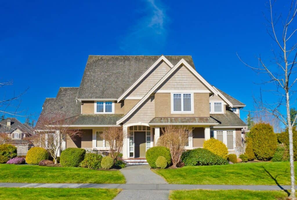 Two-story suburban house with gabled roof, beige siding, and manicured lawn. Surrounded by shrubs and small trees under a clear blue sky.