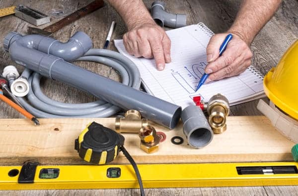Plumber's hands sketching on paper, surrounded by PVC pipes, fittings, a yellow hard hat, measuring tape, and a level on a wooden surface.