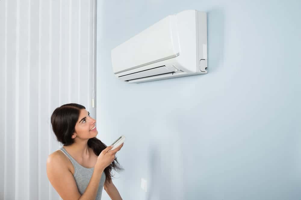 A woman in a gray tank top holds a remote control and looks at a wall-mounted air conditioner in a room with vertical blinds.