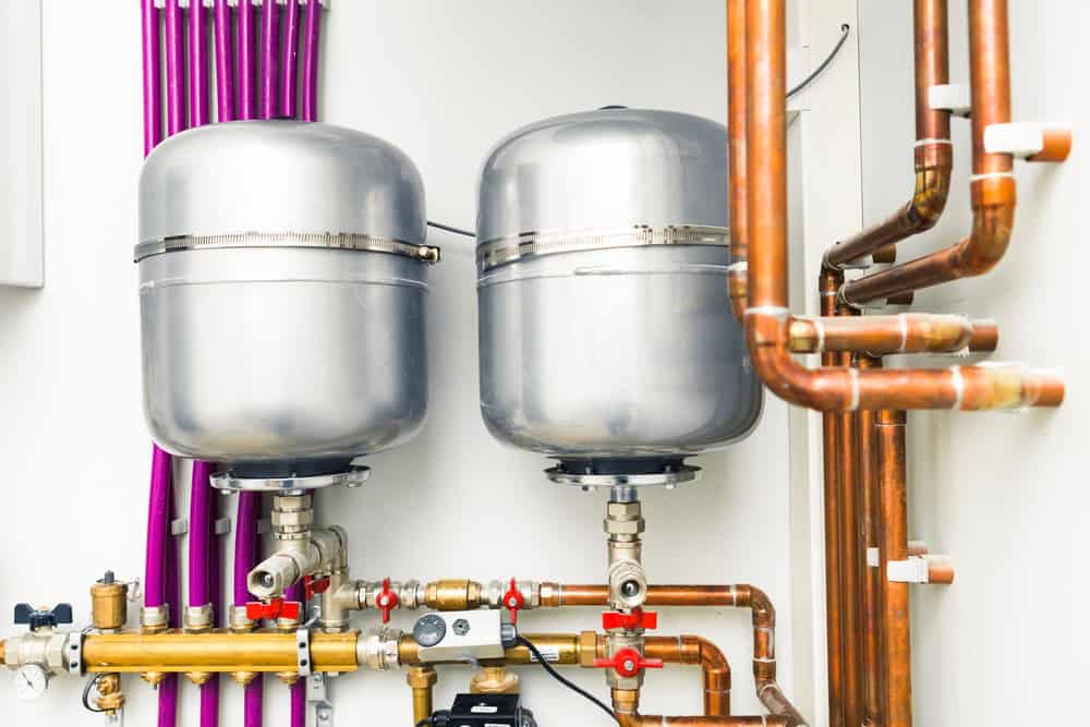 Two large silver expansion tanks with connected copper and purple pipes are mounted on a wall in a utility room.