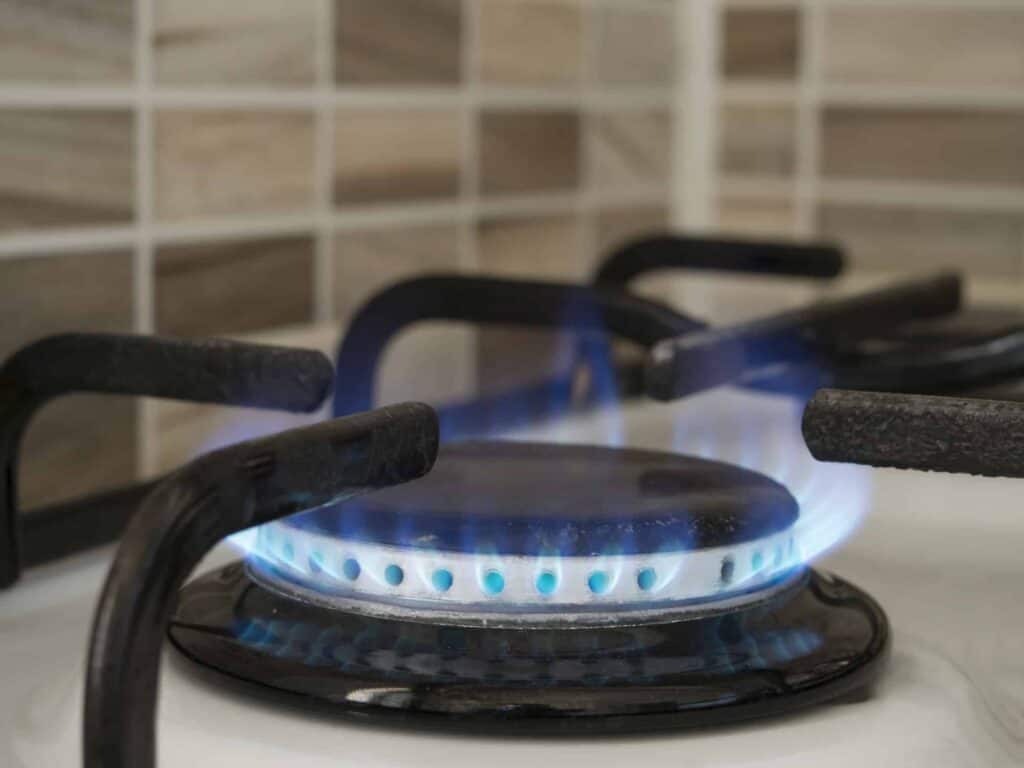 Close-up of a lit gas stove burner with visible blue flame, set against a tiled backsplash.