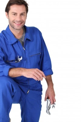 A person in a blue work jumpsuit holds a wrench, smiling at the camera.