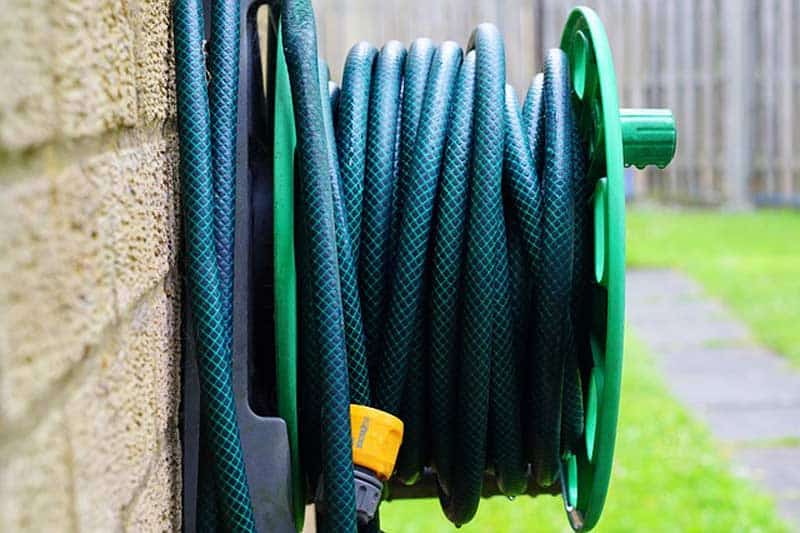 A coiled green garden hose is mounted on a wall reel, with a yellow nozzle attached, near a stone wall and a grassy area.
