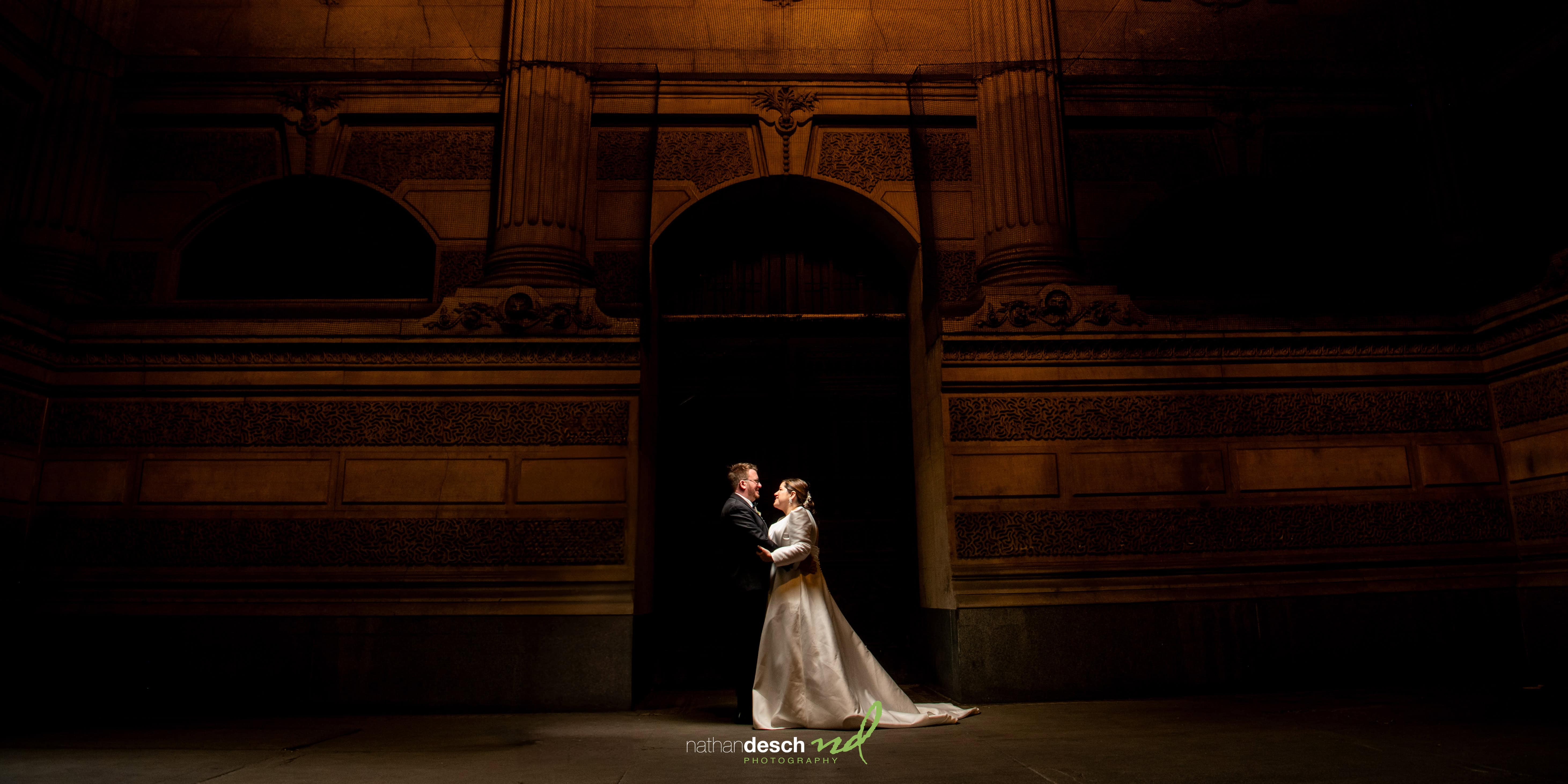 Bride and groom embrace under dramatic lighting outside Tendenza at night, showcasing elegant wedding attire and architecture.