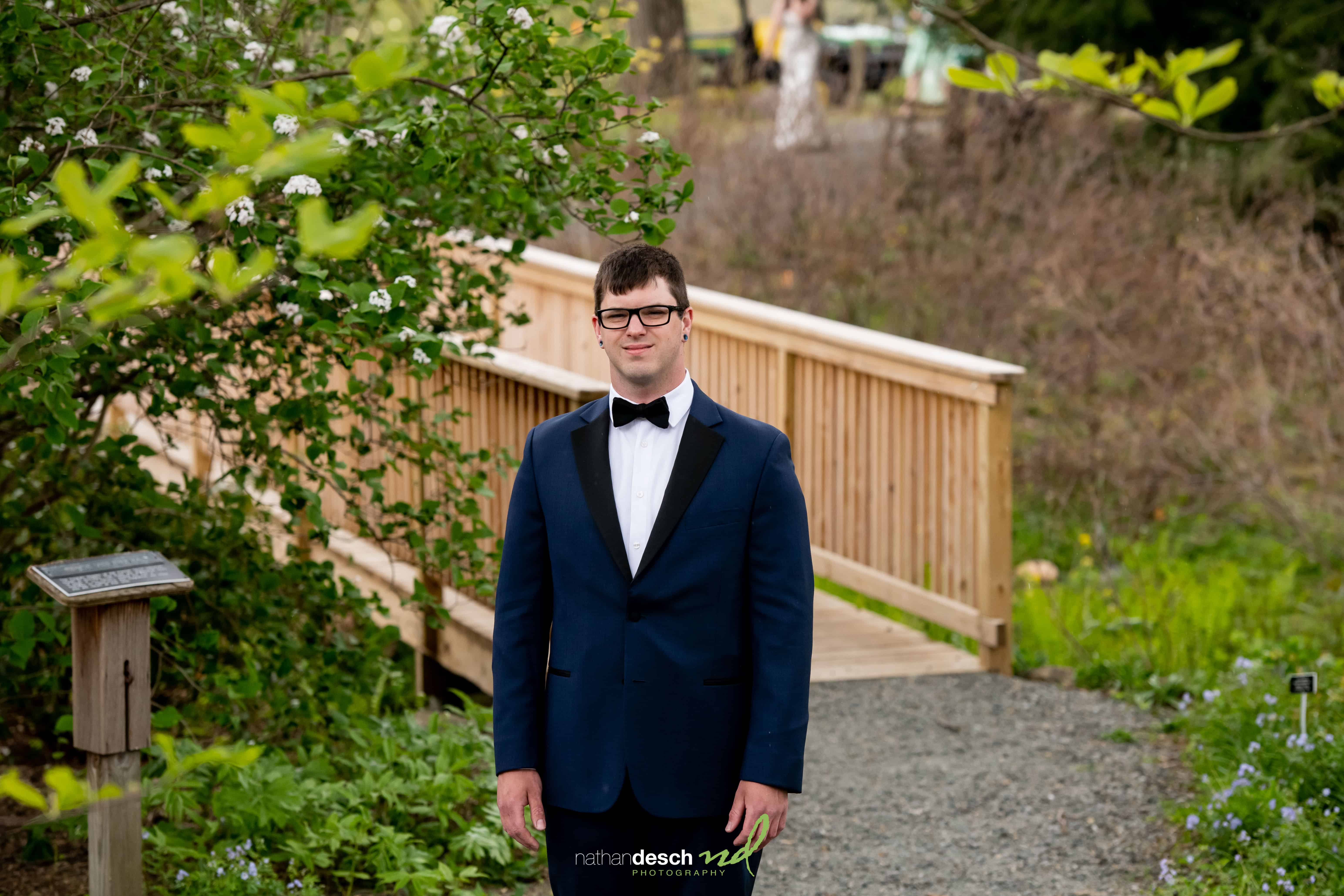 Groom in navy tuxedo stands on gravel path by wooden bridge at Frelinghuysen Arboretum wedding, lush greenery and flowers.