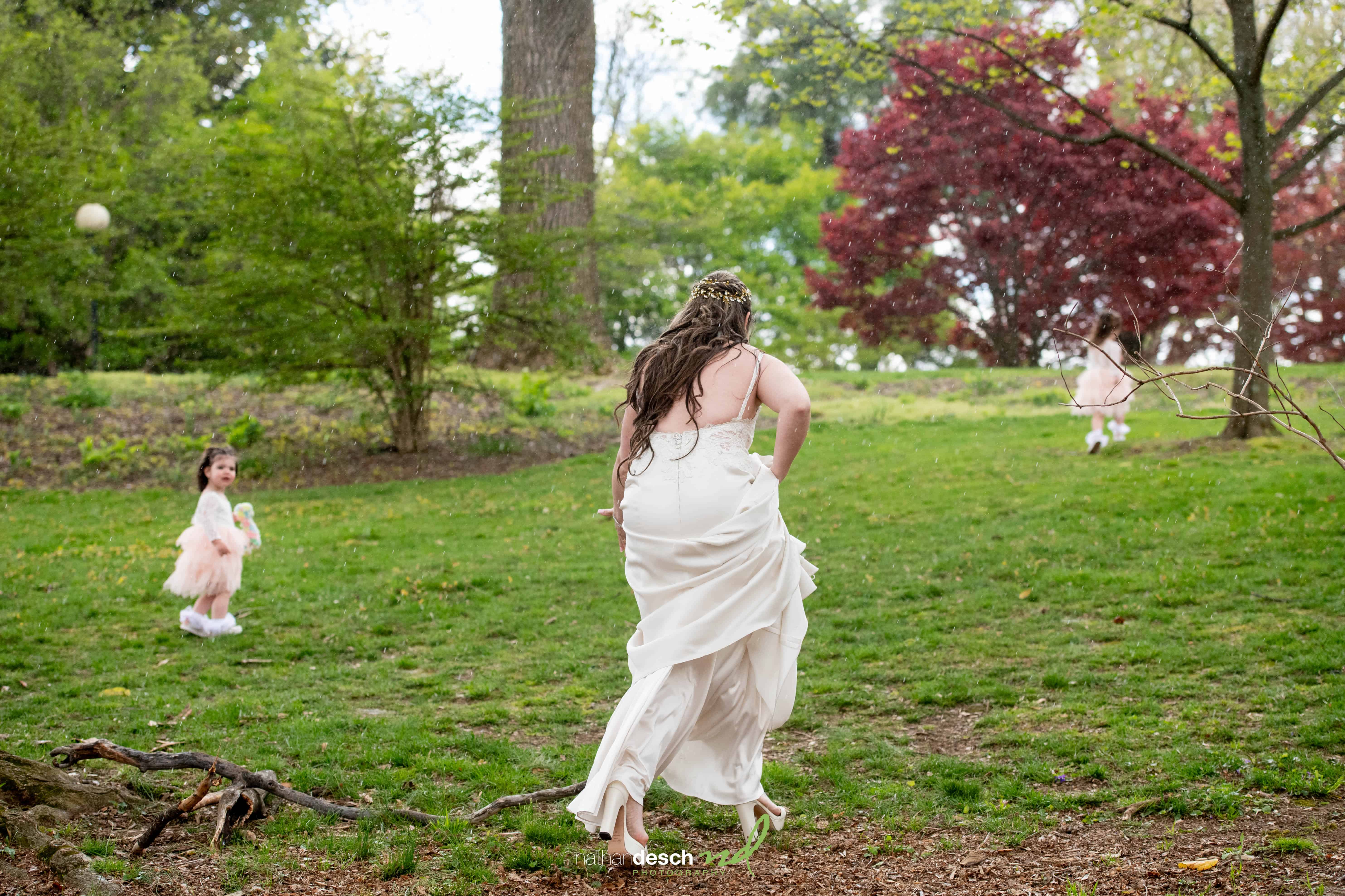 Bride in white dress runs across Frelinghuysen Arboretum lawn toward two flower girls, surrounded by lush green trees.