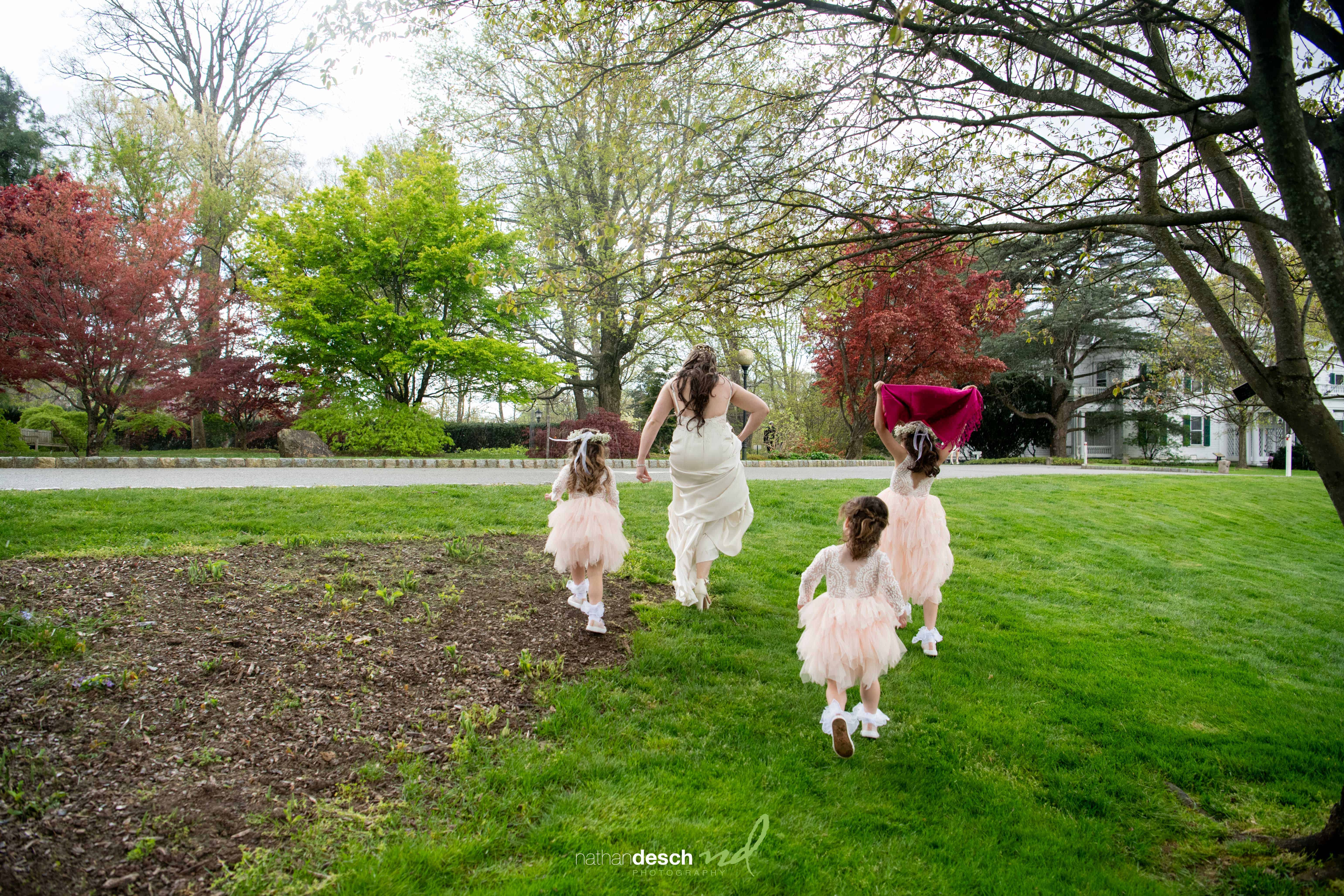 Bride in white gown and three flower girls in pink dresses run on grass at Frelinghuysen Arboretum spring wedding.