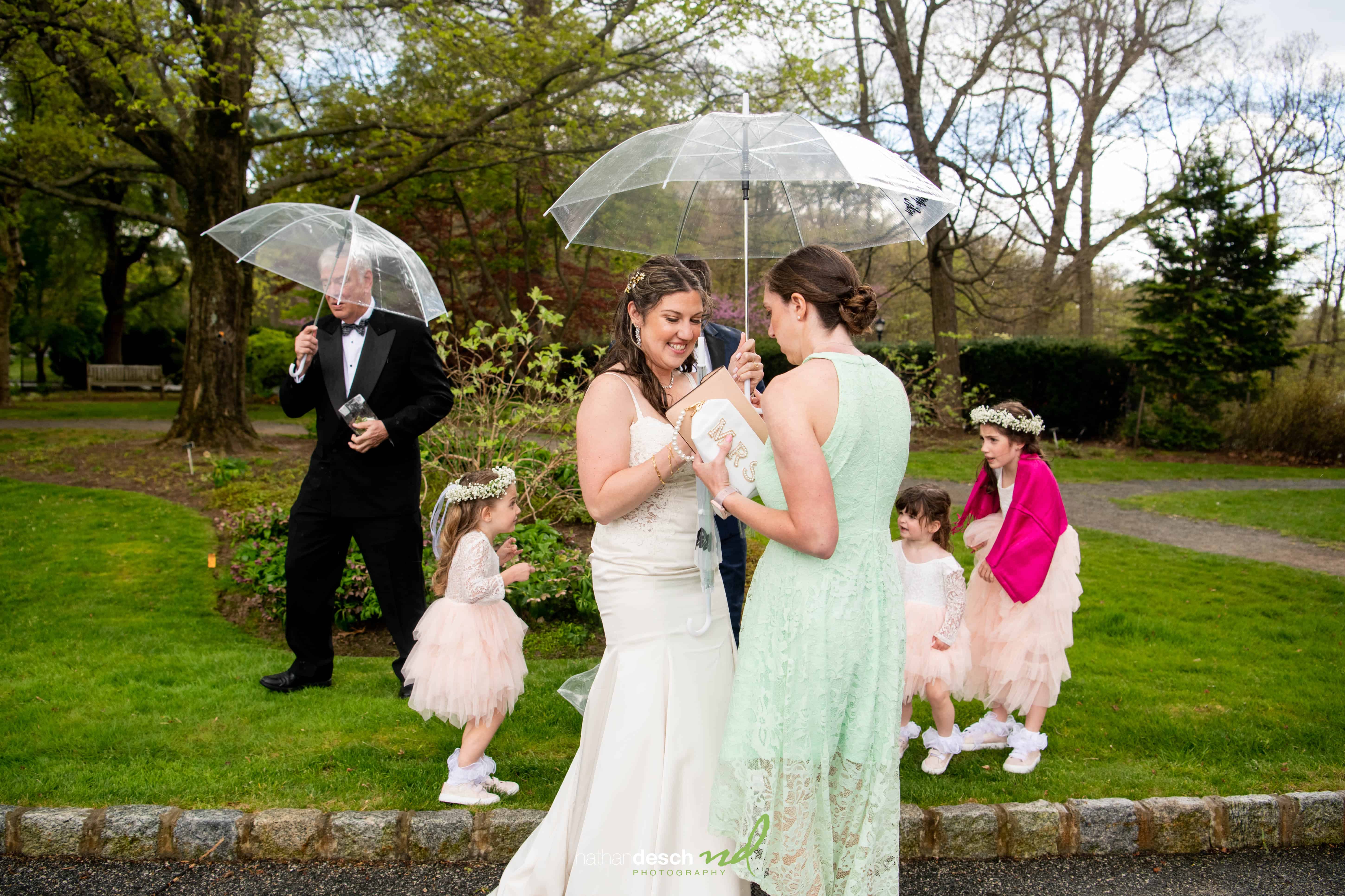 Two brides exchange vows under a clear umbrella in the rain at Frelinghuysen Arboretum, with flower girls by their side.