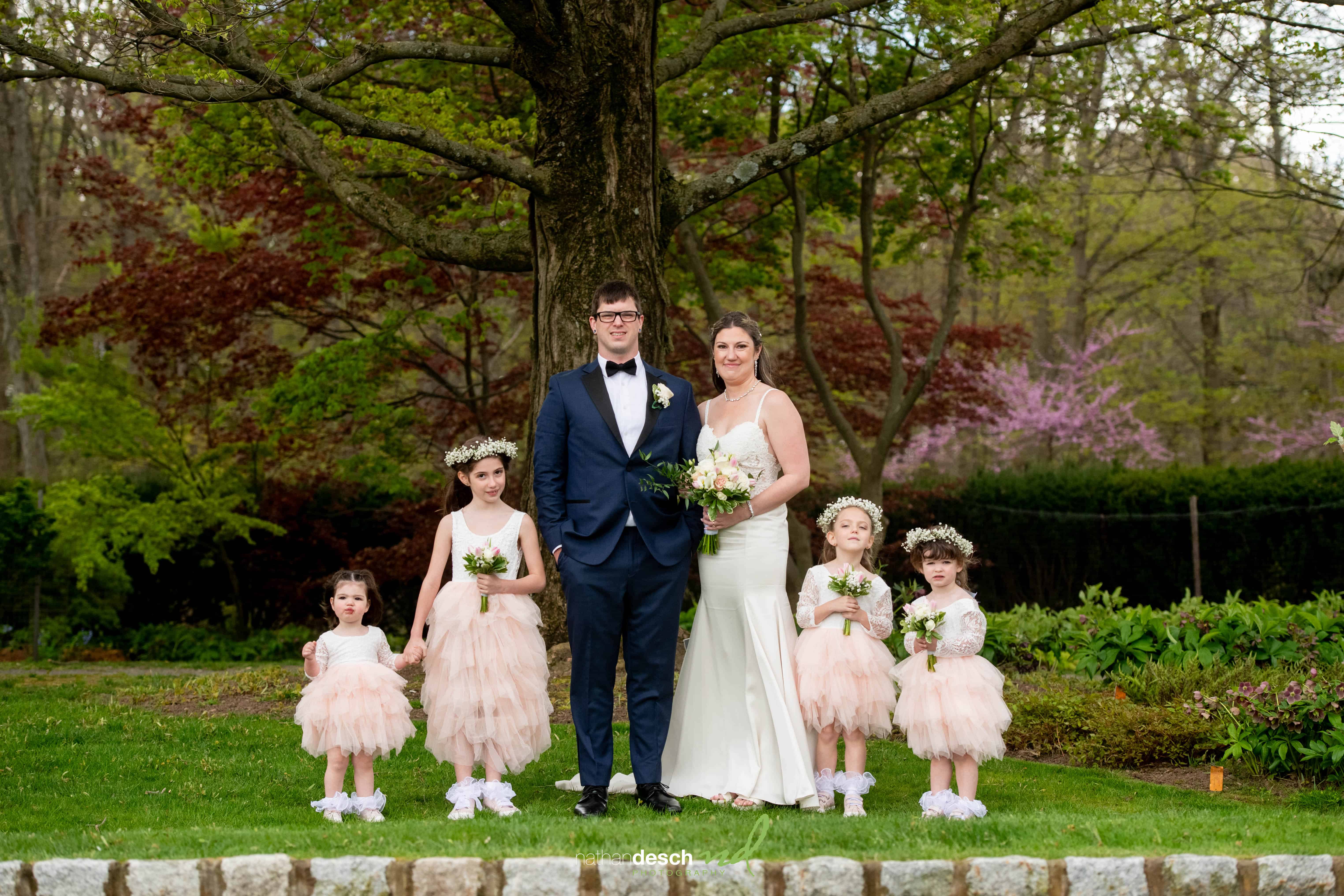 Bride and groom with four flower girls in pink dresses at Frelinghuysen Arboretum spring wedding, under blooming trees.