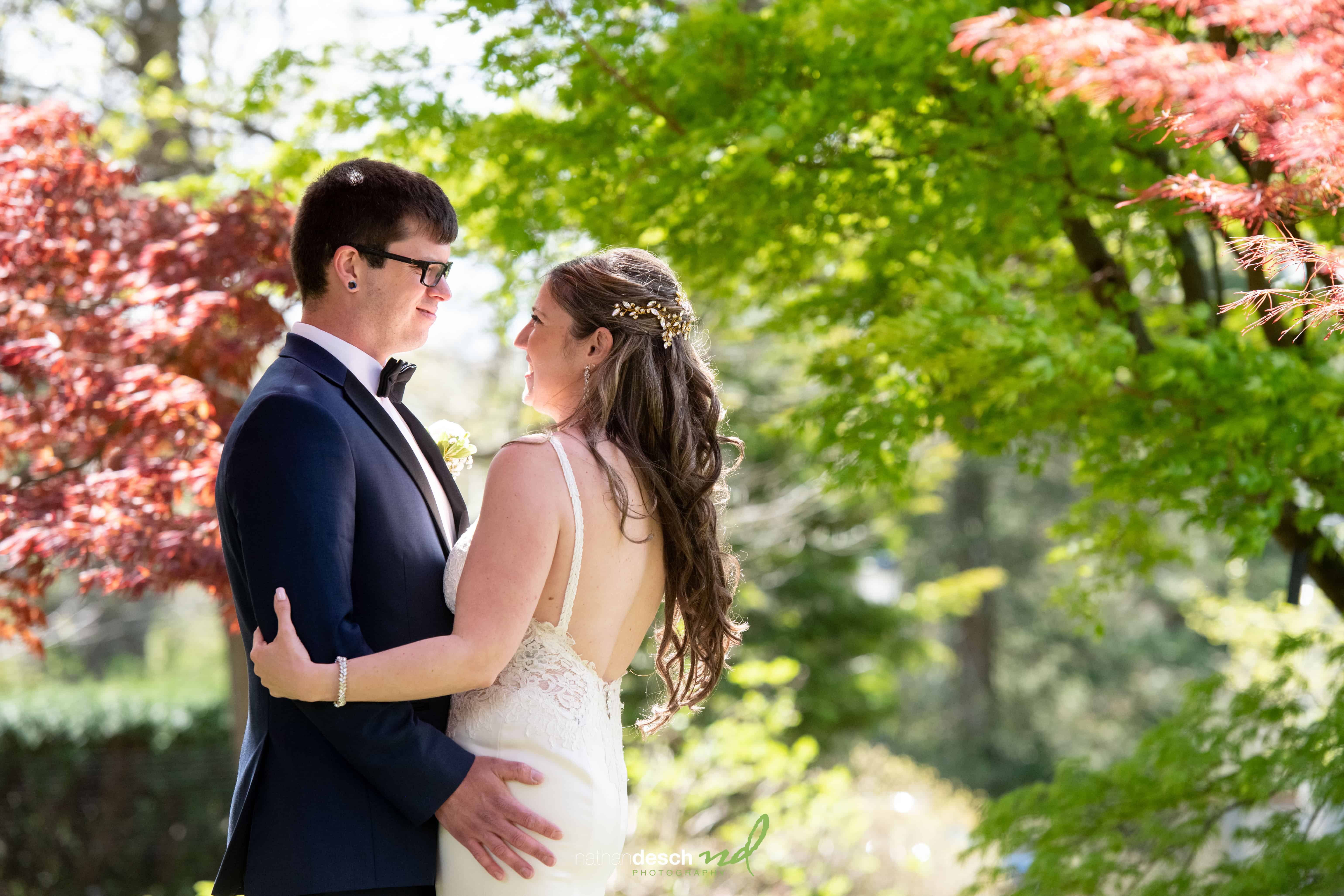 Bride and groom smile, gazing at each other outdoors among lush green and red trees on a sunny wedding day.