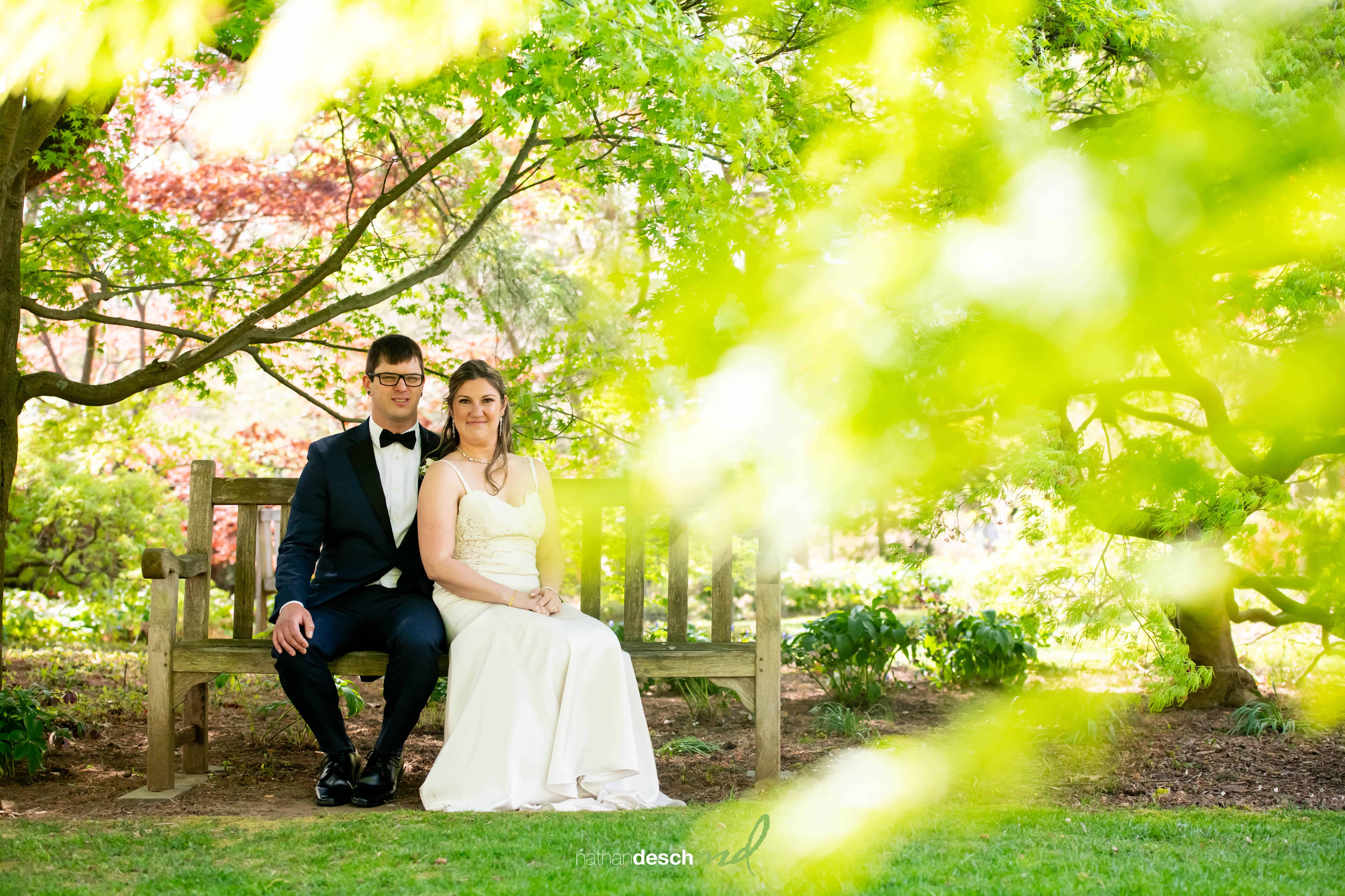 Bride and groom smiling on a wooden bench in a sunlit garden, surrounded by lush greenery and wedding attire.