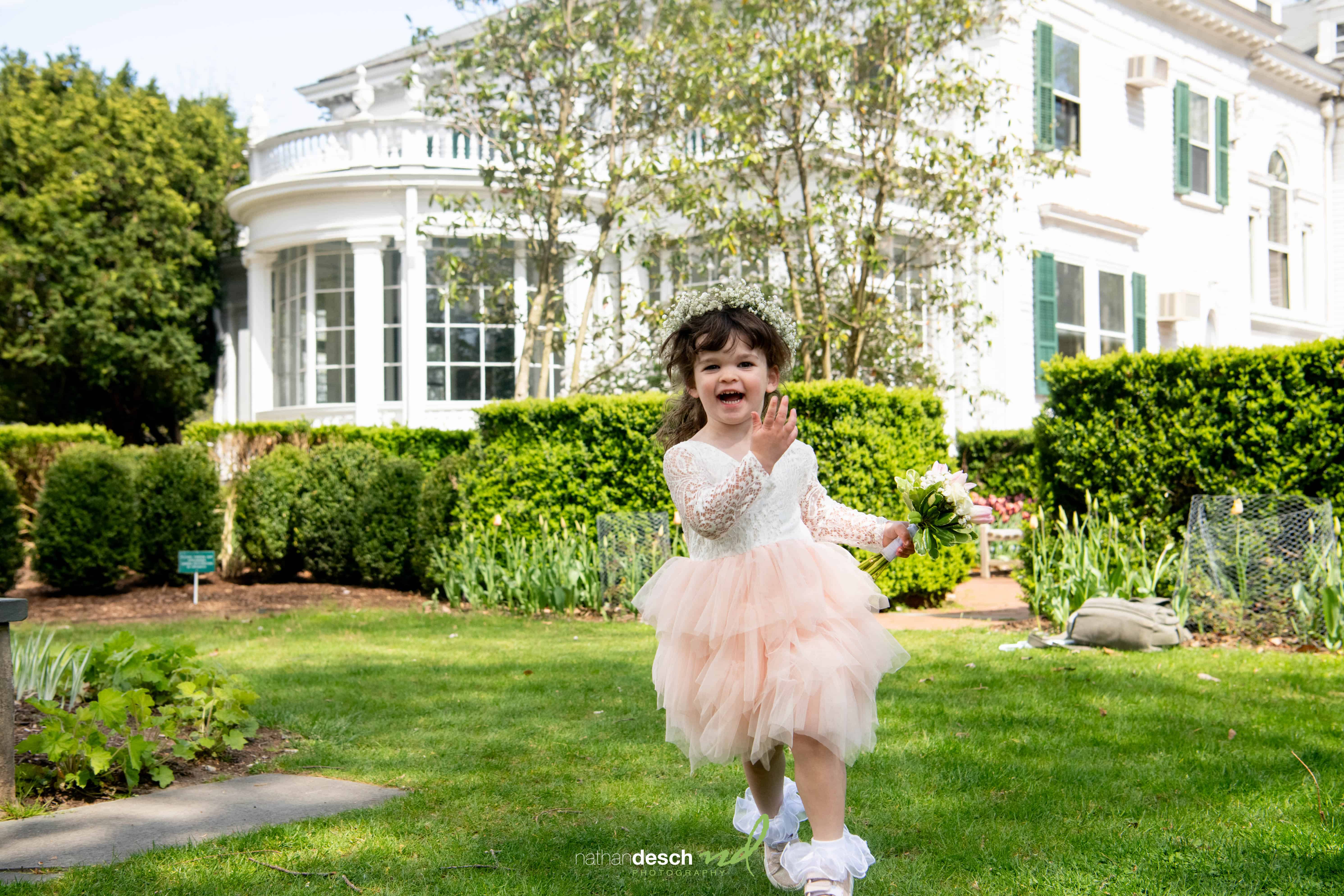 Smiling flower girl in peach tulle dress waves on lawn at Frelinghuysen Arboretum wedding, white house and greenery behind.