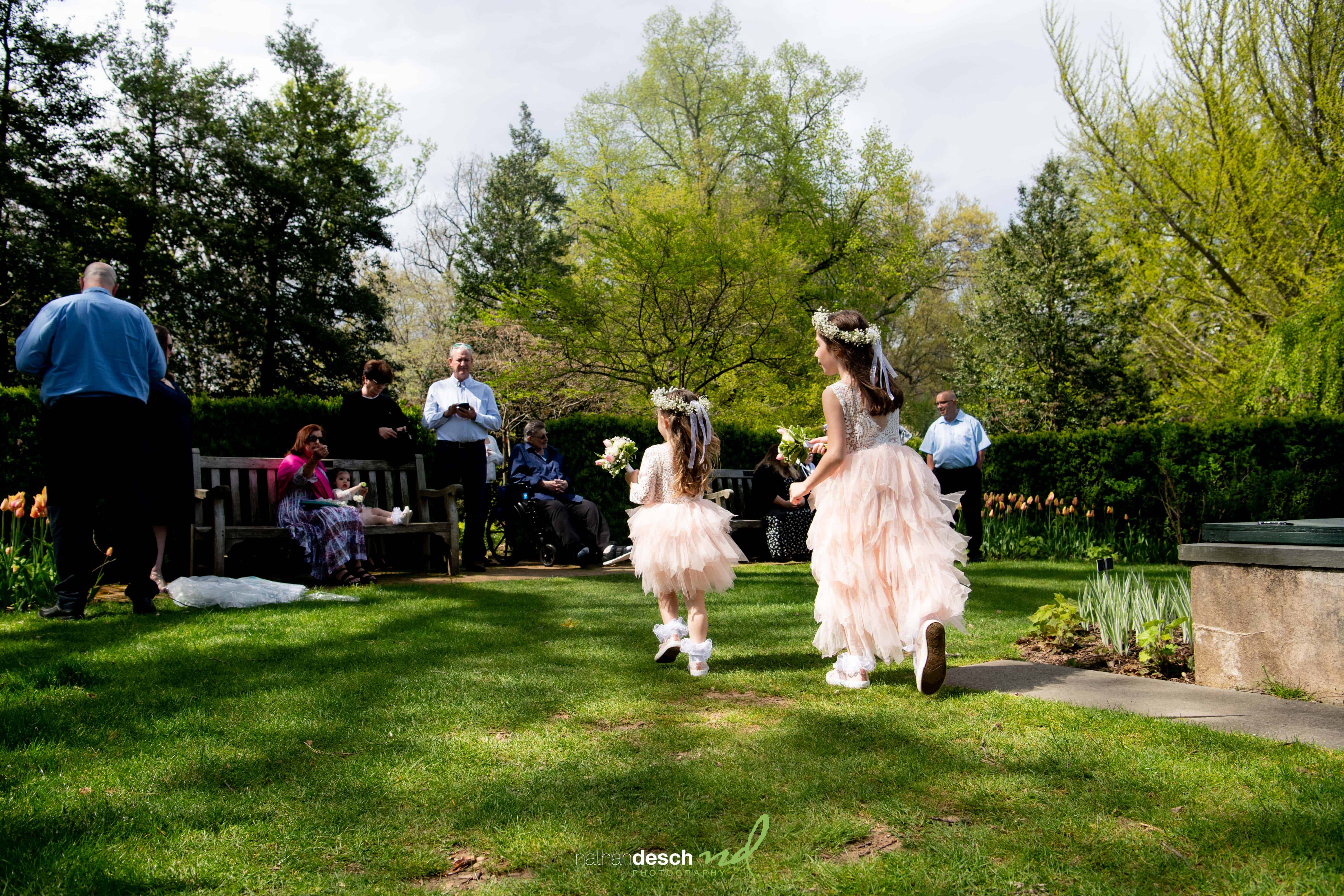 Two young flower girls in pink ruffled dresses and flower crowns walk across a sunny garden wedding toward guests.