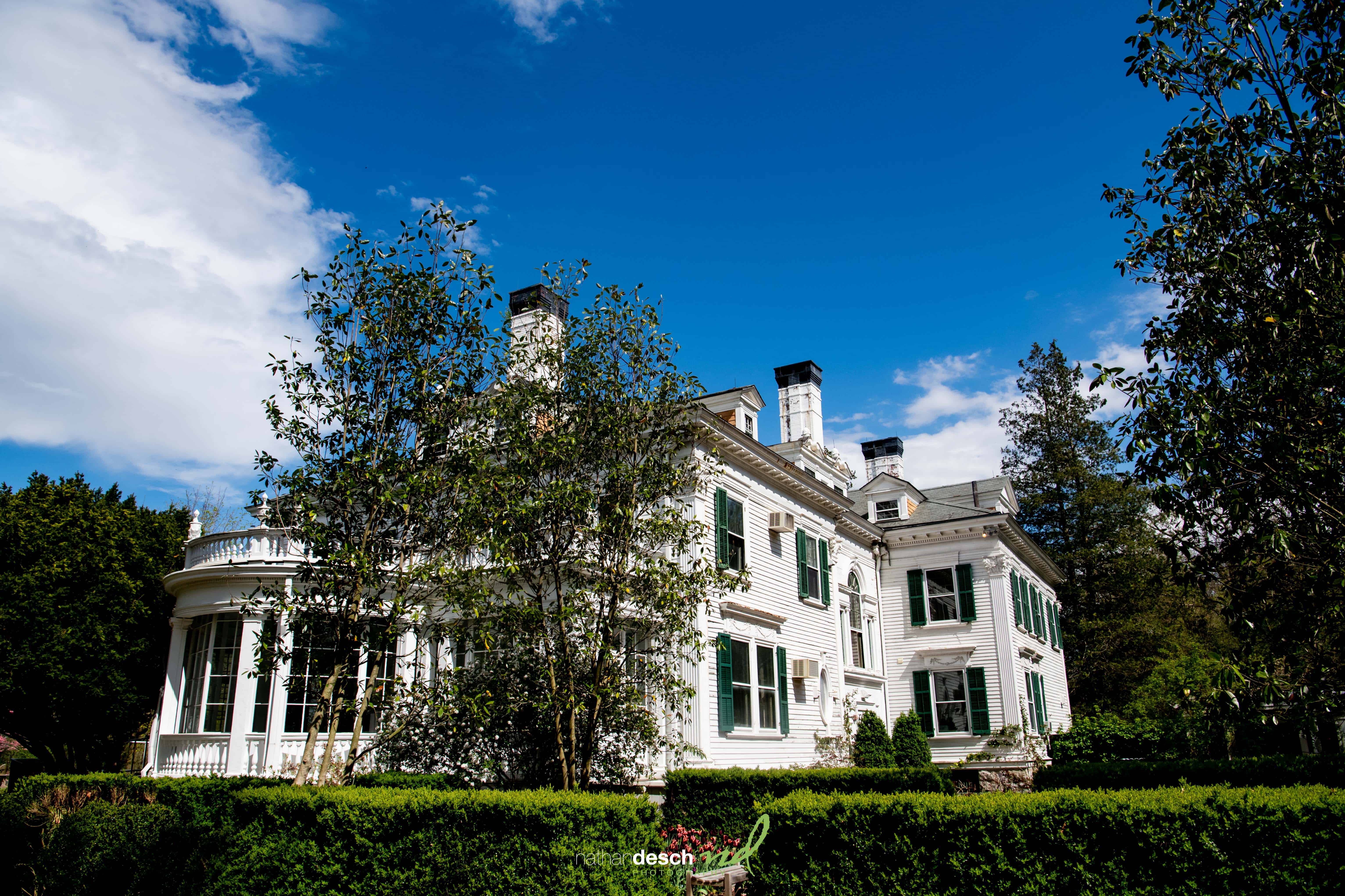 Elegant white historic house with green shutters, chimneys, and lush greenery under a blue sky—Billy and Kaitlyn’s wedding venue.