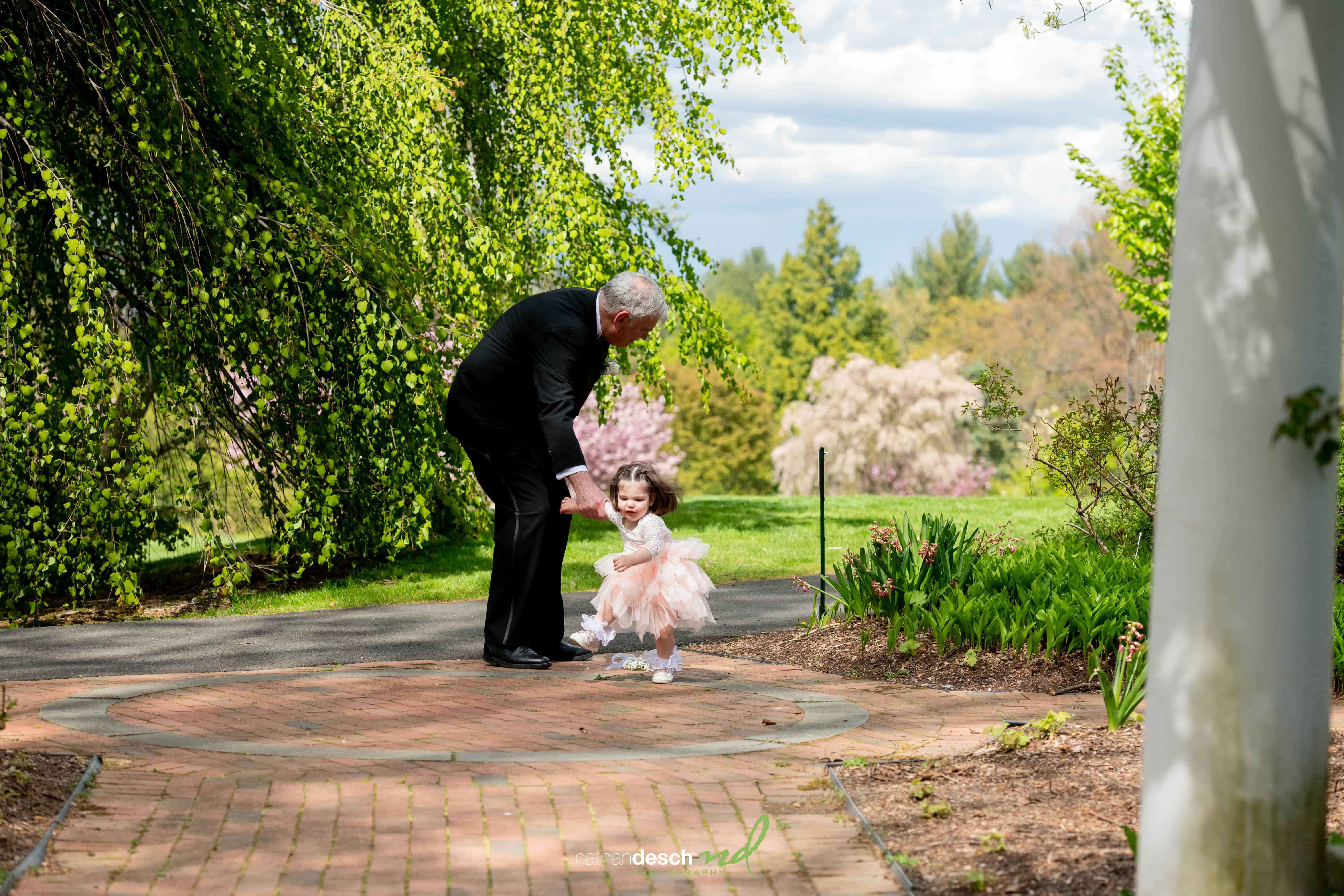 Older man in black suit helps smiling toddler in pink dress walk on brick path with lush greenery and blooming trees behind.