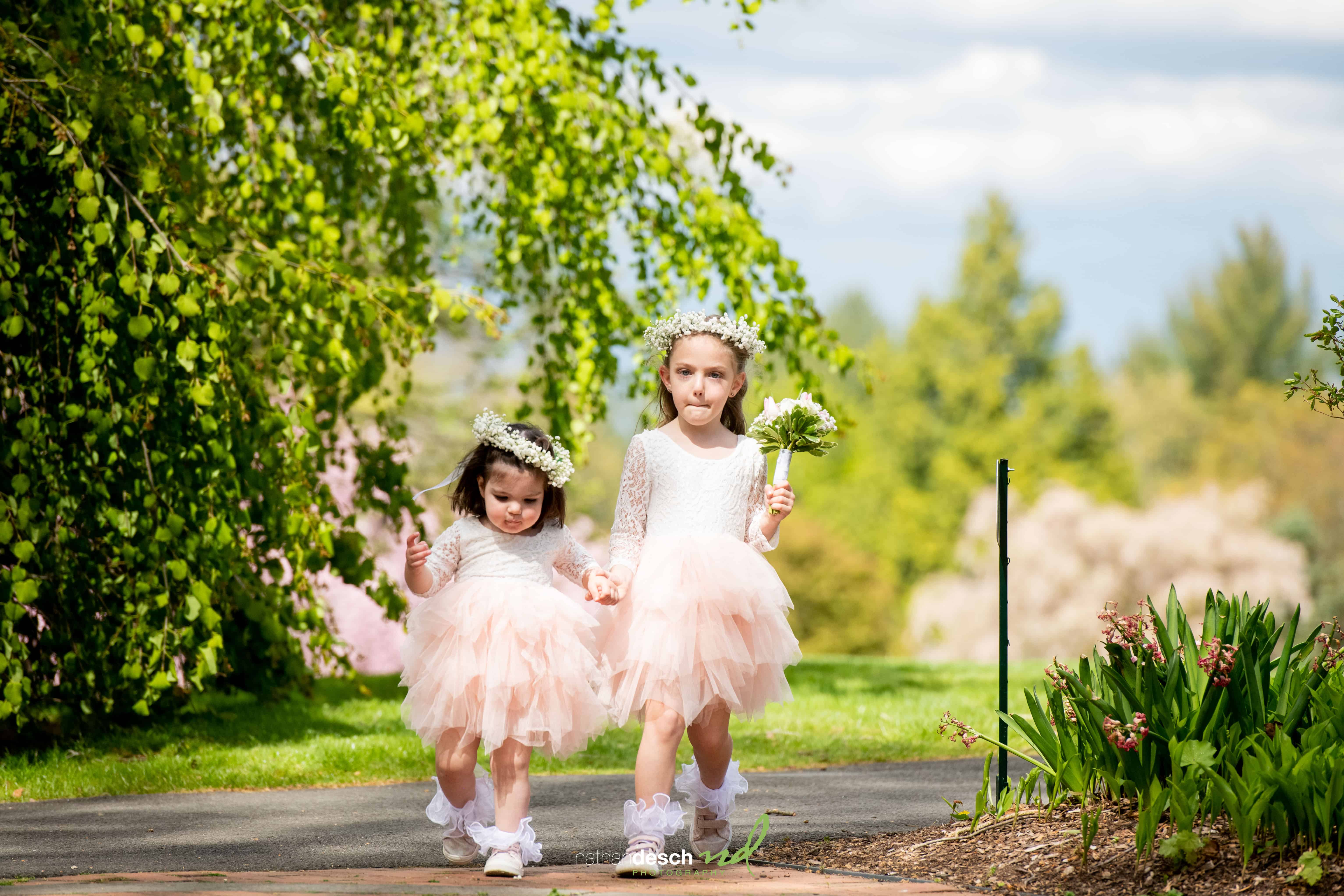 Two flower girls in pink tulle dresses and crowns walk hand-in-hand on a sunlit garden path with lush greenery.