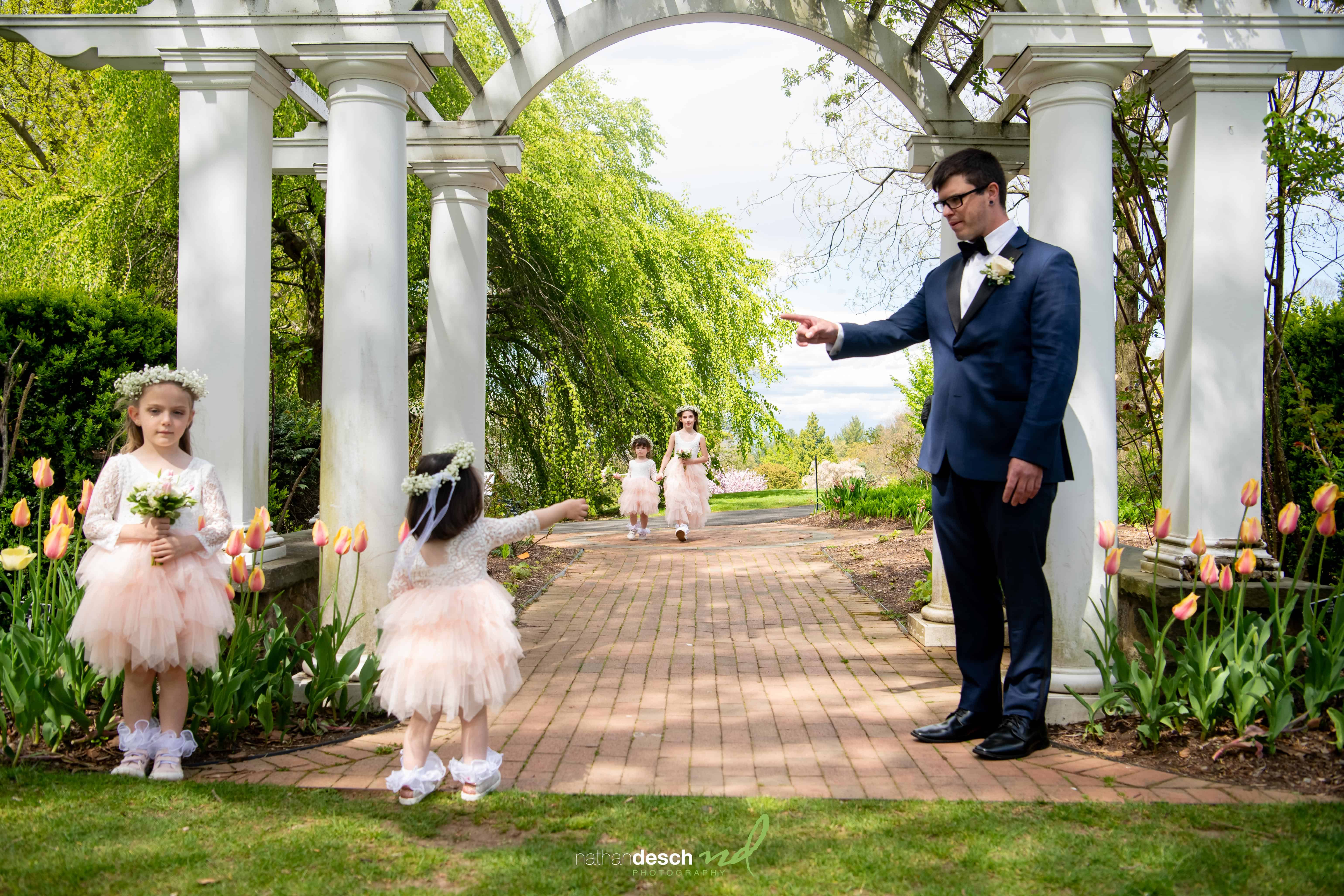 Groom and flower girls smile in garden, bride walking under white pergola with tulips during spring wedding ceremony.