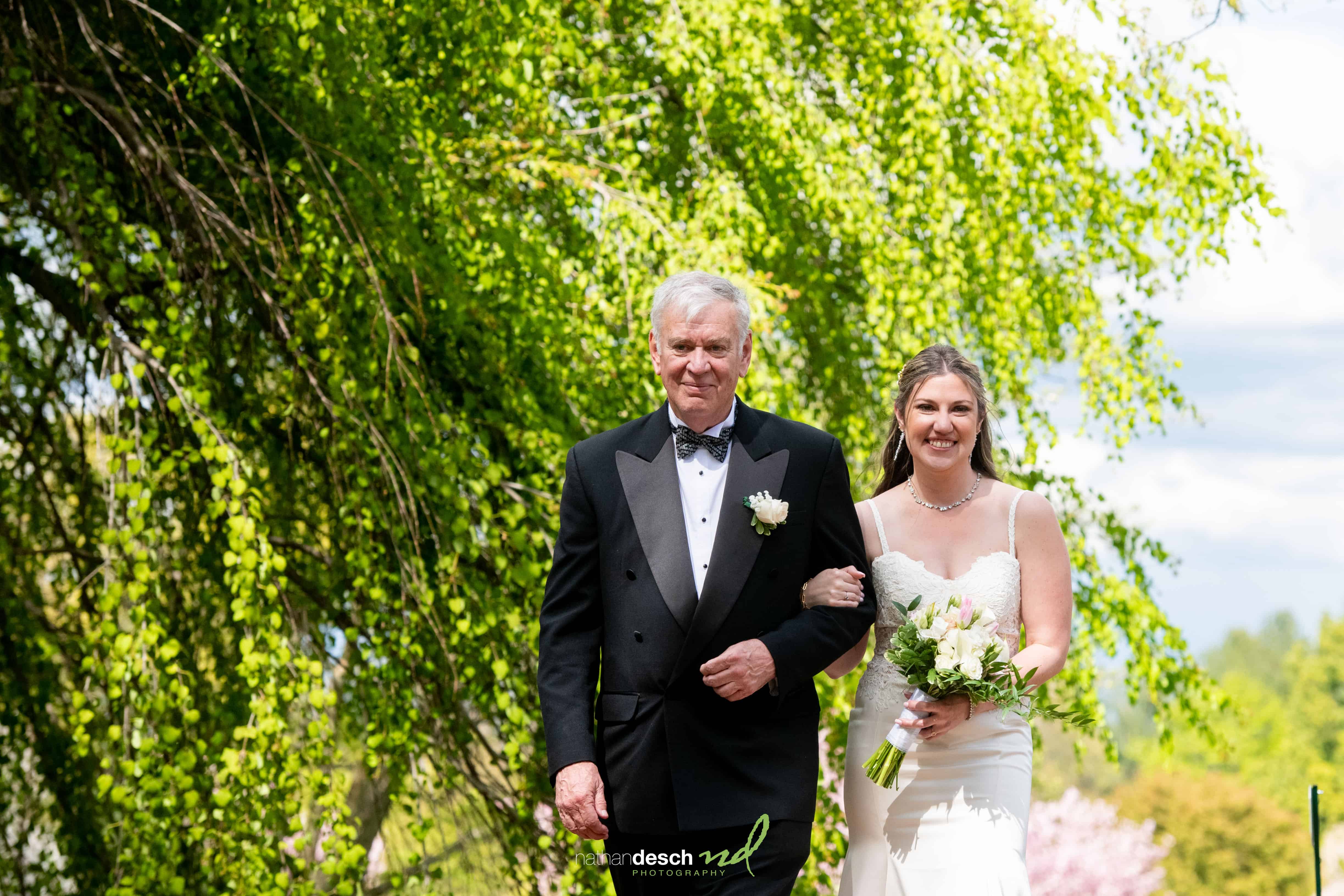 Bride and groom smiling arm in arm outdoors, bride in white dress with bouquet, groom in tux, surrounded by lush greenery.