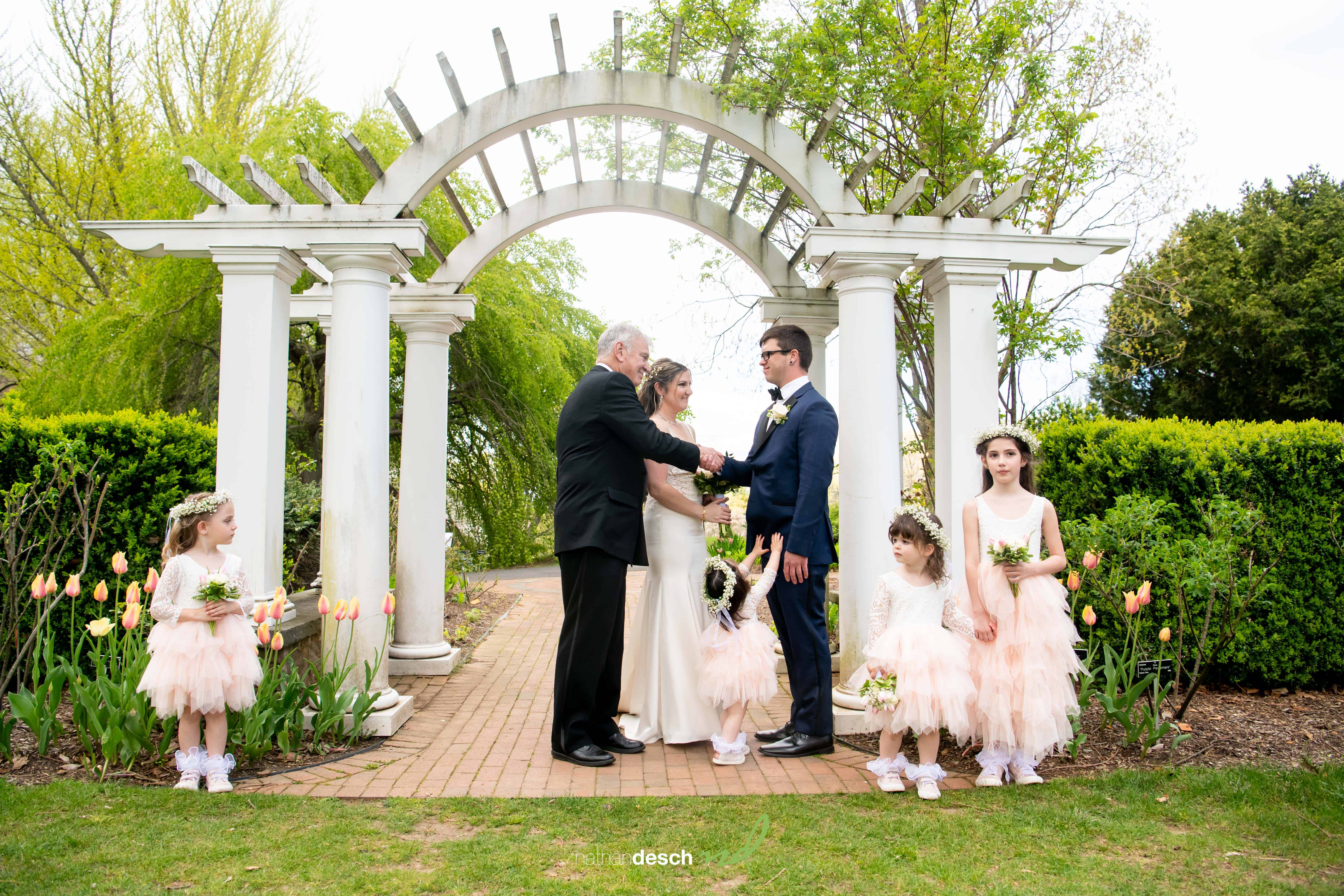 Bride and groom hold hands under a white garden arch with flower girls in peach dresses, surrounded by tulips and greenery.