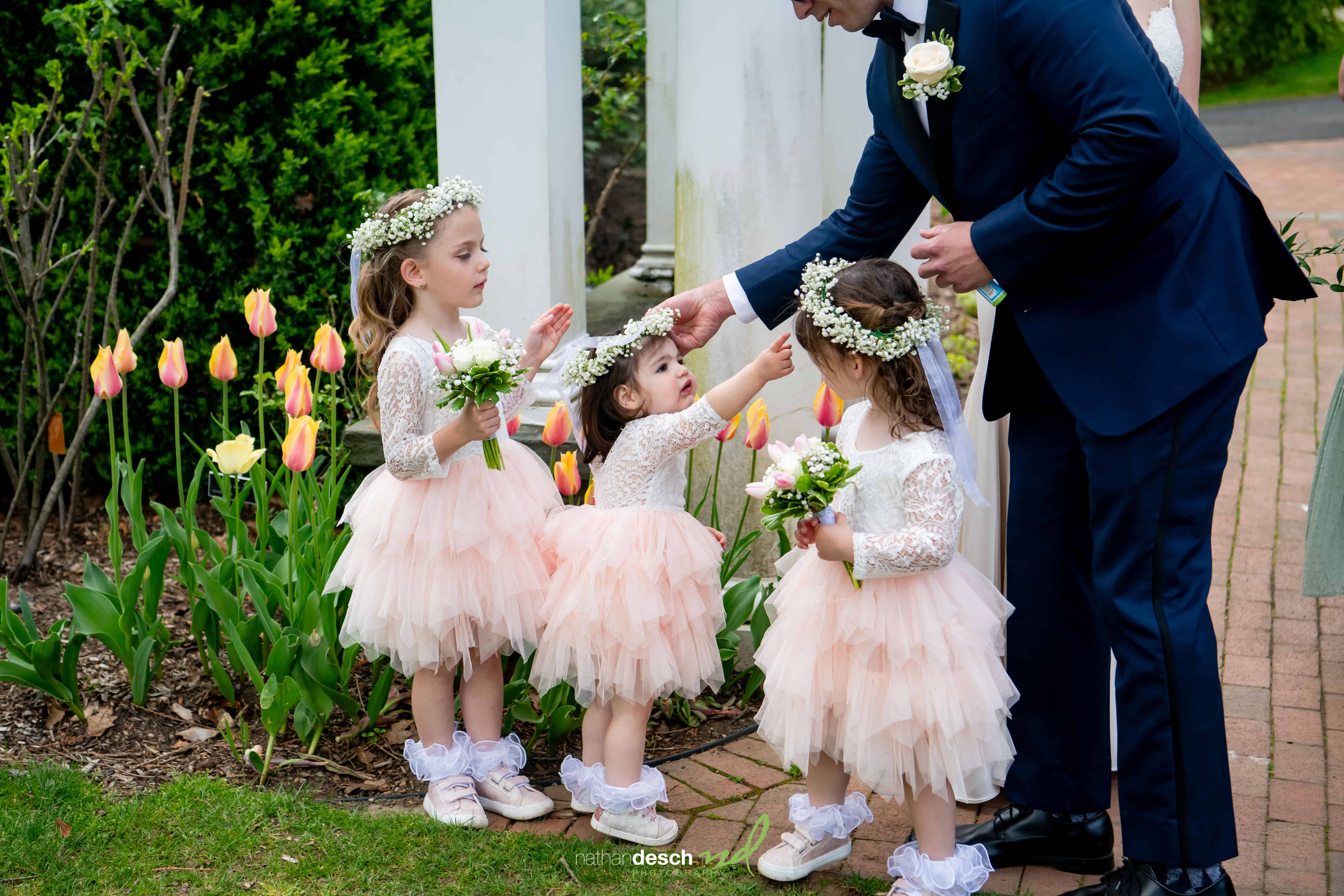 Billy in a navy suit greets three flower girls in peach dresses with floral headbands, surrounded by blooming tulips.
