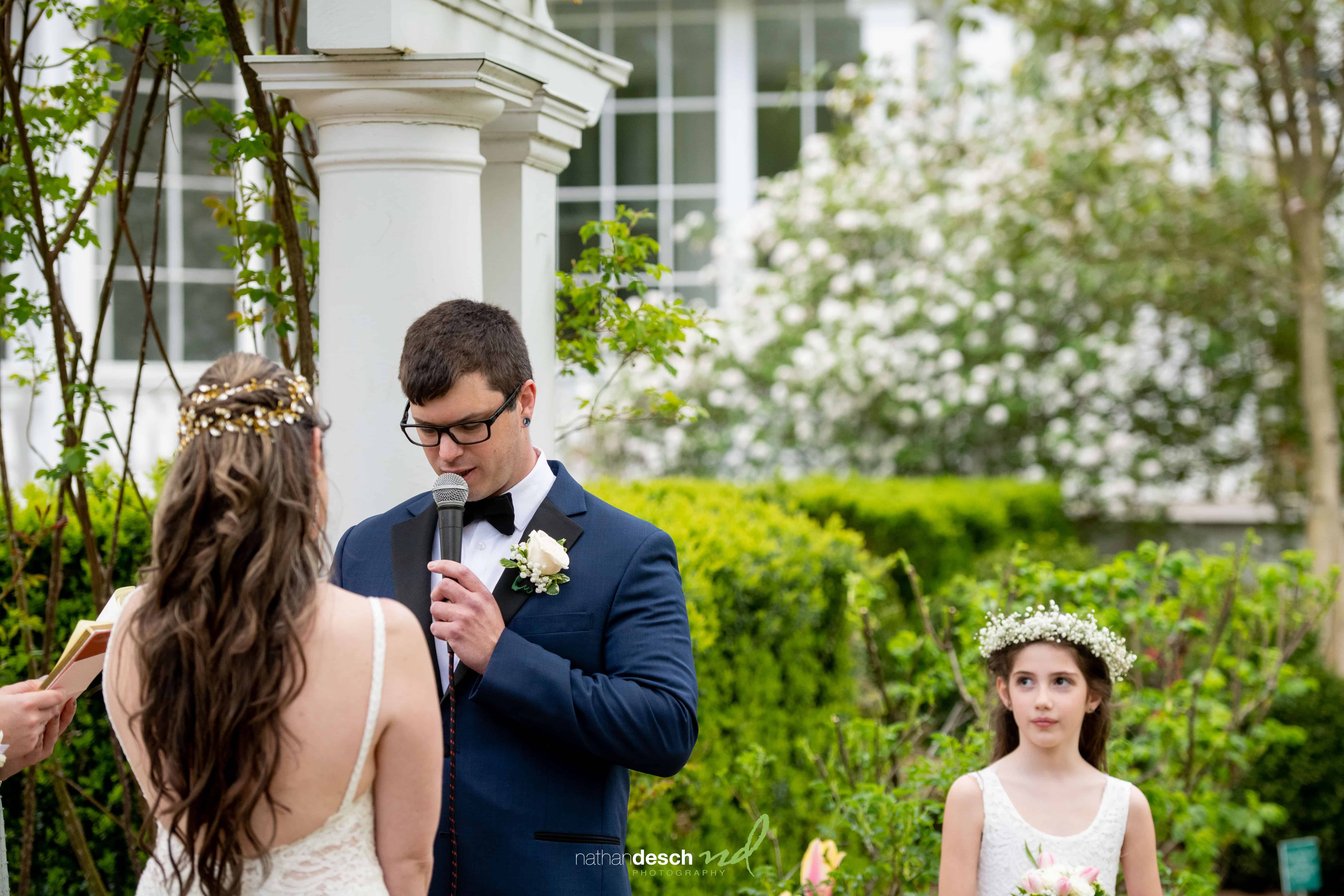 Groom reads vows beside bride in white dress and flower girl with crown at Kaitlyn and Billy’s outdoor wedding ceremony.