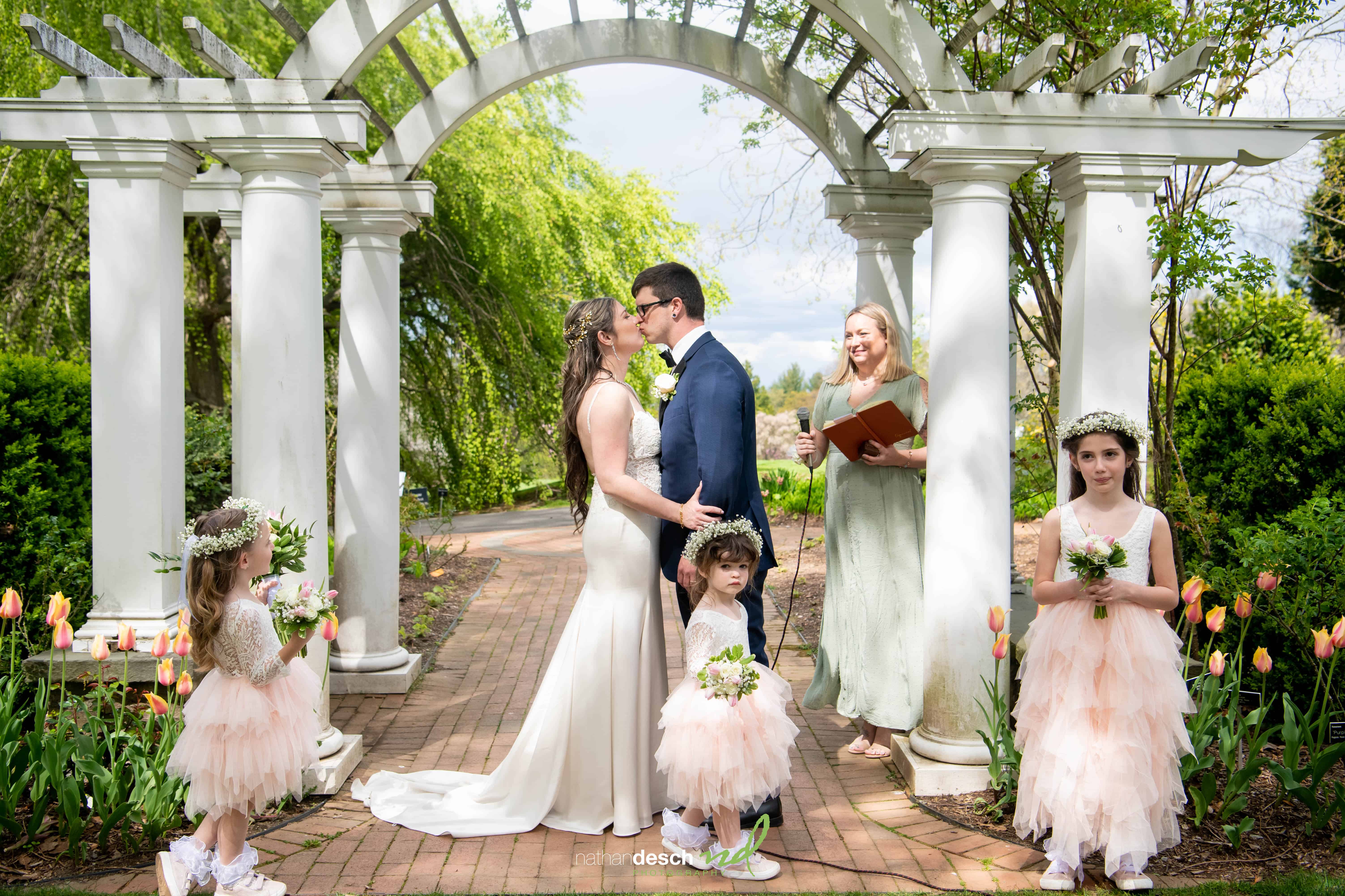 Kaitlyn and Billy share a kiss under a white garden arch, surrounded by bridesmaids, flower girls, tulips, and lush greenery.