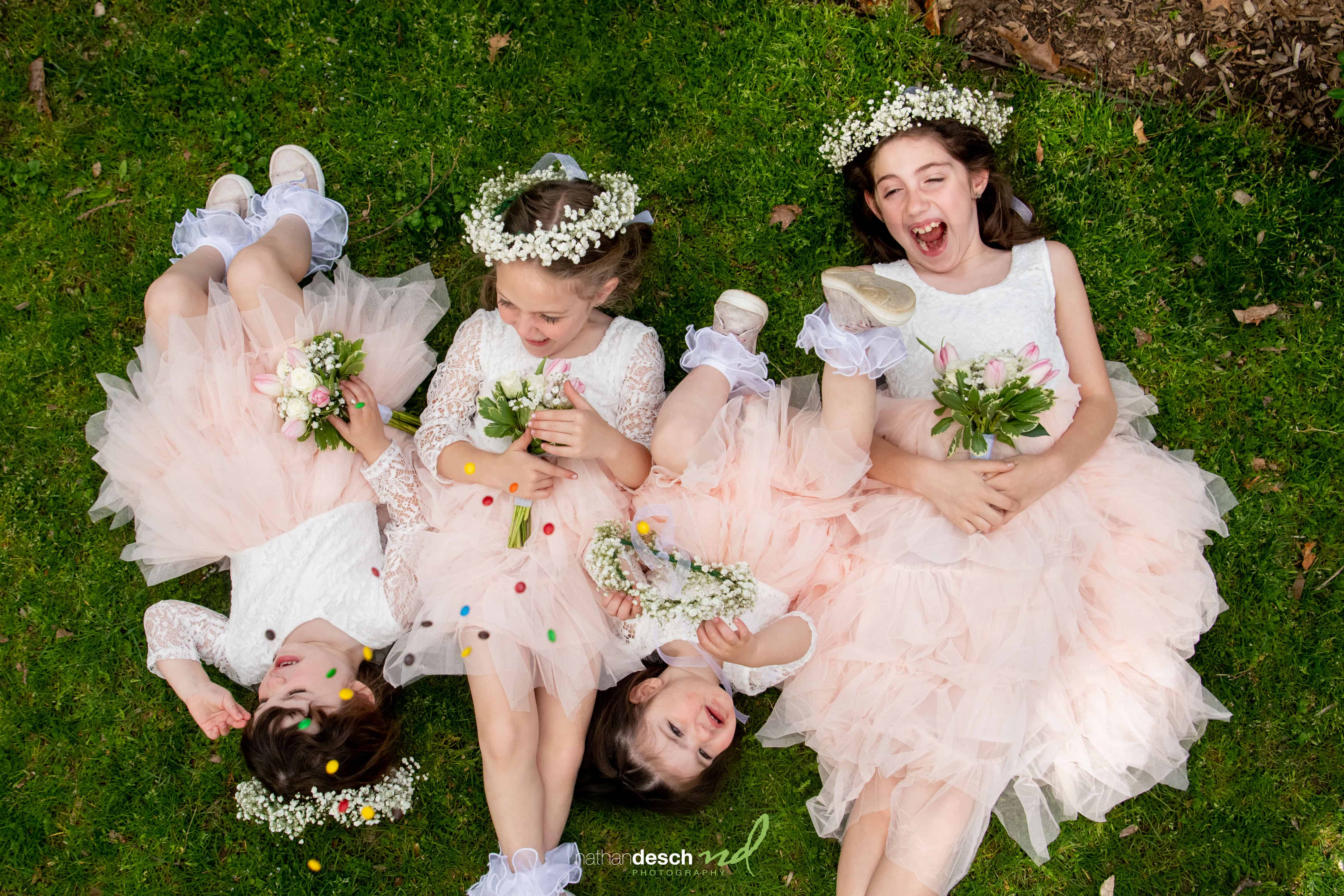 Four flower girls in white lace dresses with bouquets and candy, laughing together on green grass at a joyful outdoor wedding.