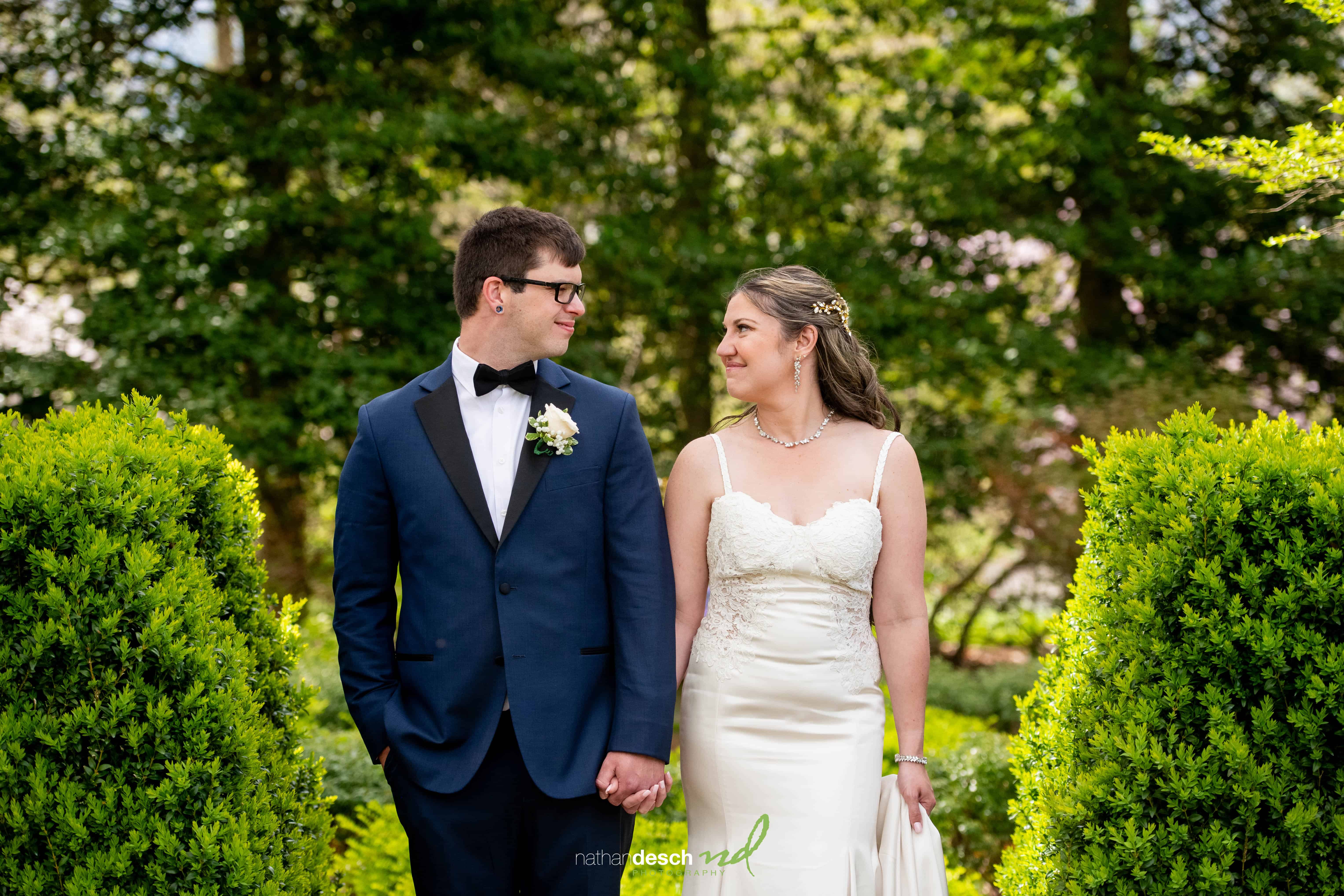 Bride and groom hold hands, smiling at each other in a lush green outdoor garden on their wedding day.