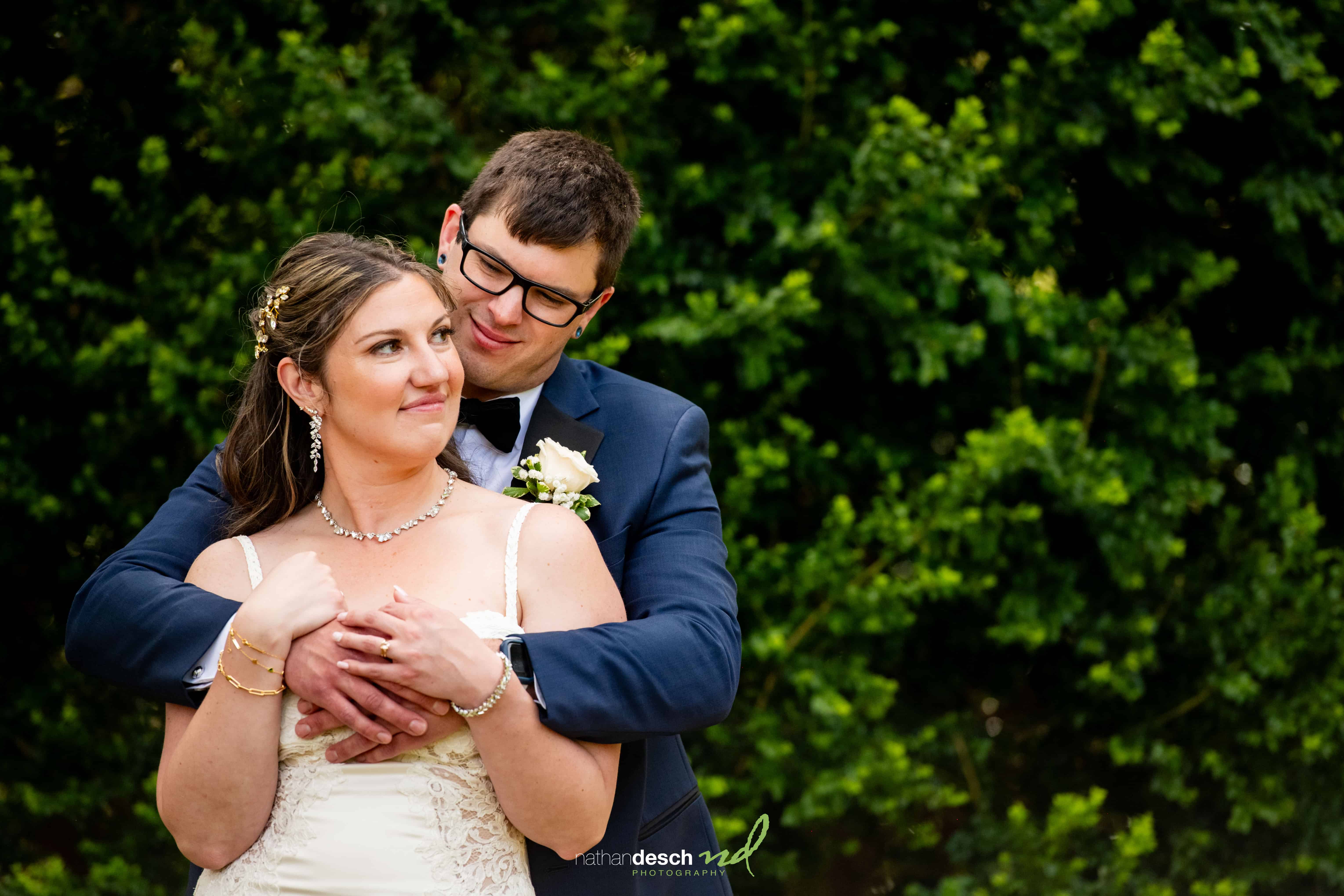 Groom hugs smiling bride from behind outdoors, surrounded by lush green foliage on their wedding day.