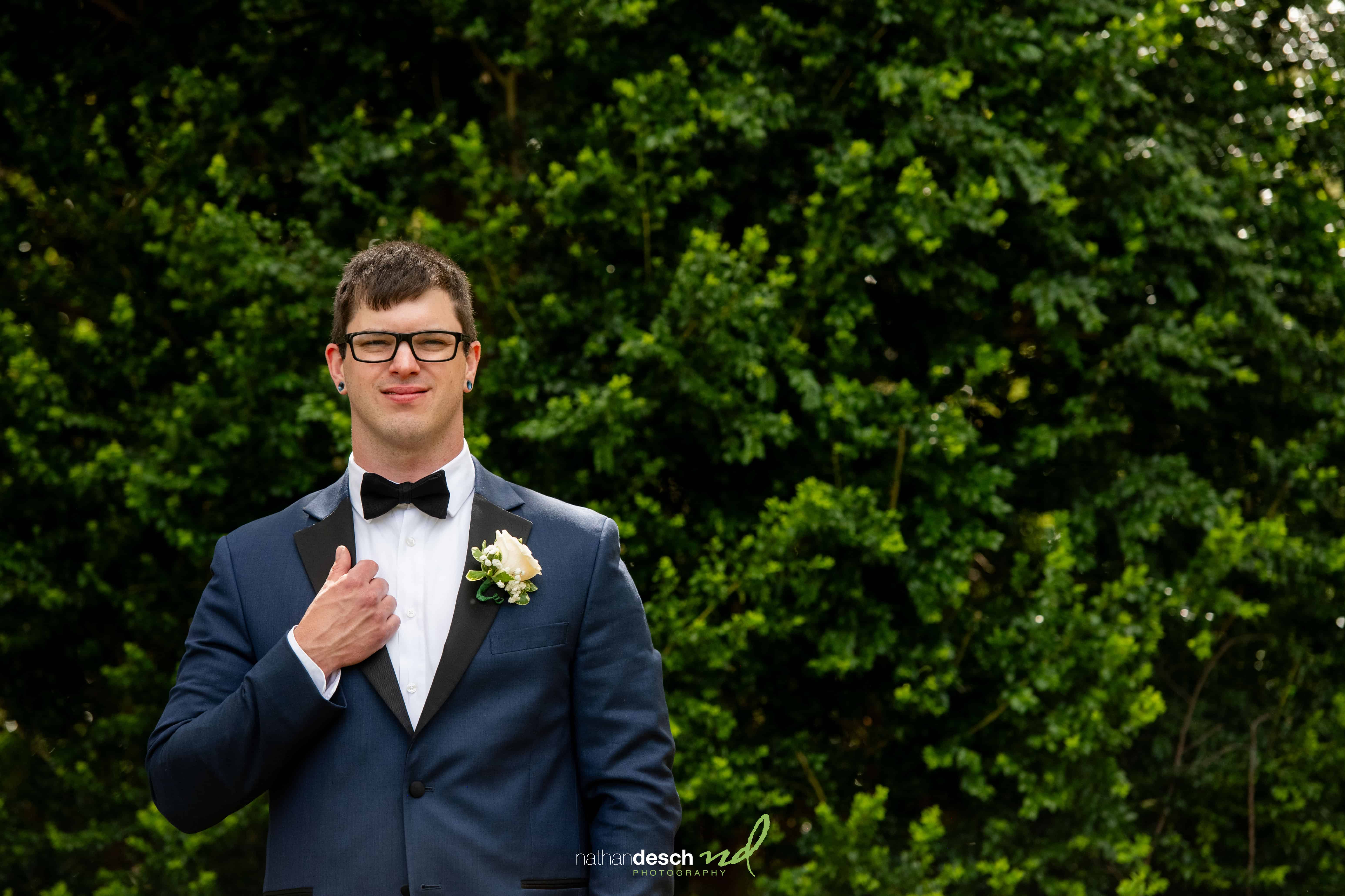 Groom in navy tuxedo with bow tie and boutonniere smiles outdoors against lush green foliage on wedding day.