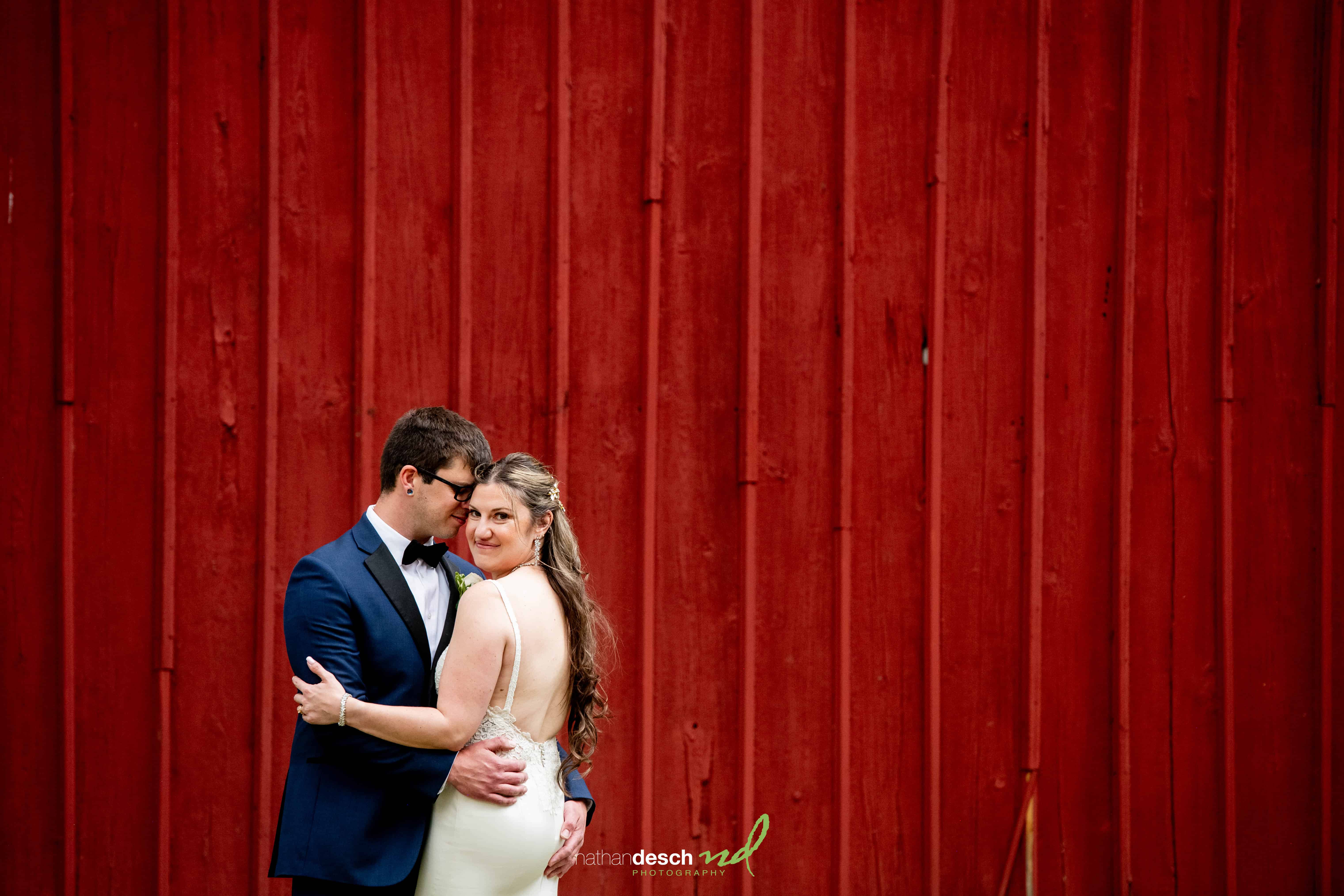 Bride and groom embrace and smile in front of rustic red wooden wall at their wedding, wearing classic attire.