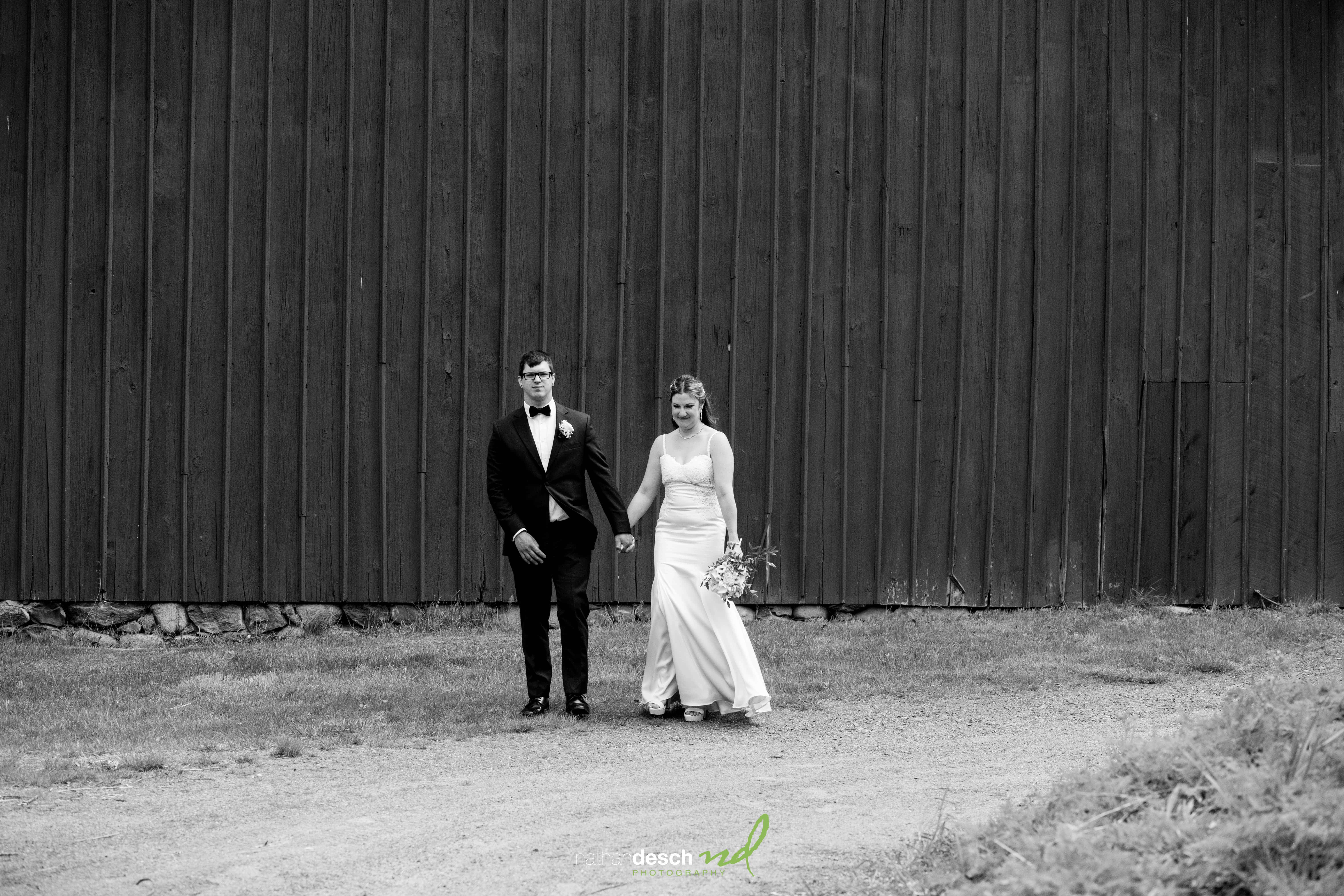 Bride and groom holding hands in front of rustic barn, black-and-white wedding photo, bride with bouquet and strapless gown.