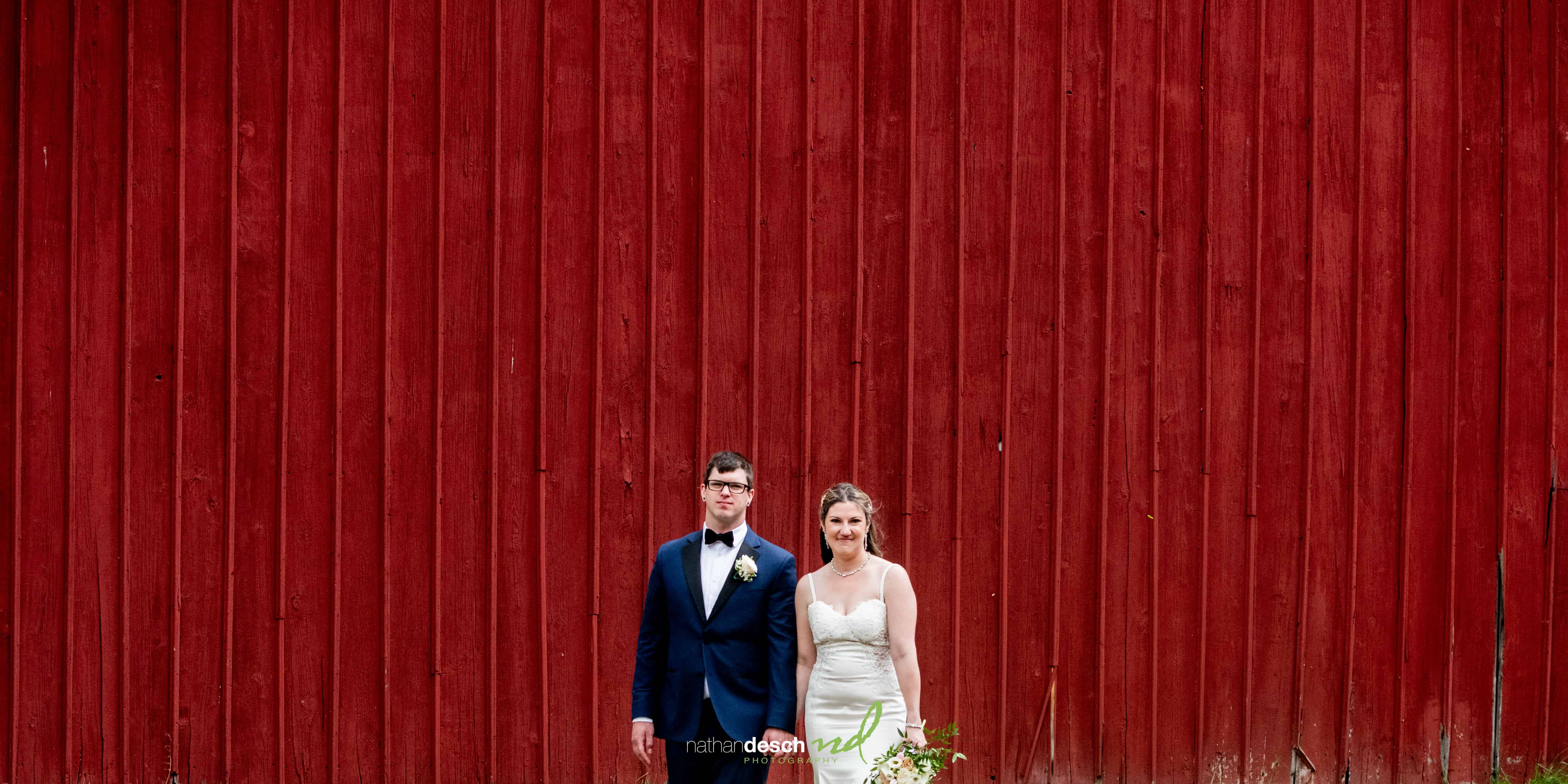 Bride in white dress and groom in navy suit pose together by rustic red wooden wall, smiling at wedding photographer.