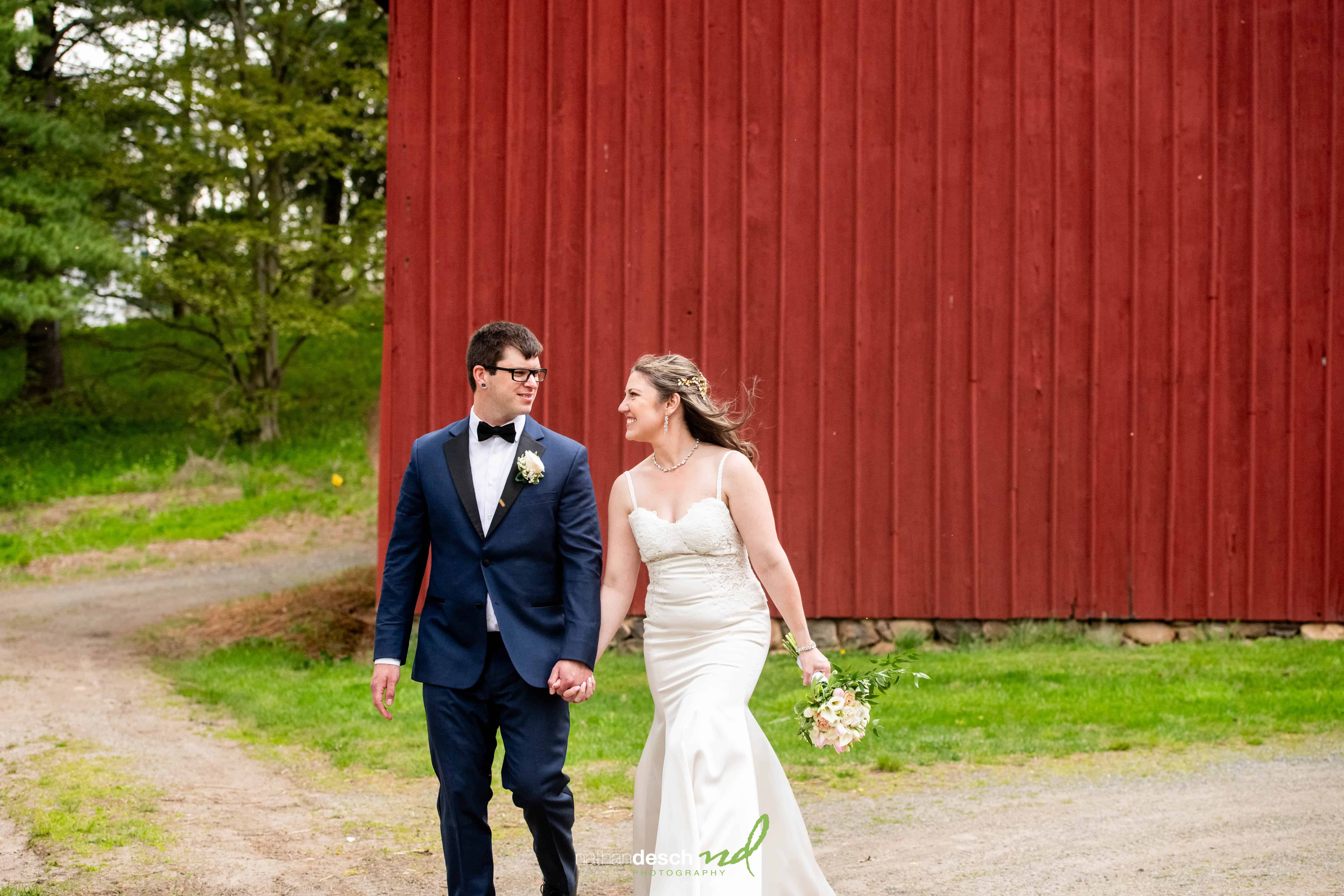 Bride and groom hold hands smiling, walking by a red barn and green trees; bride in white dress, bouquet; groom in navy suit.