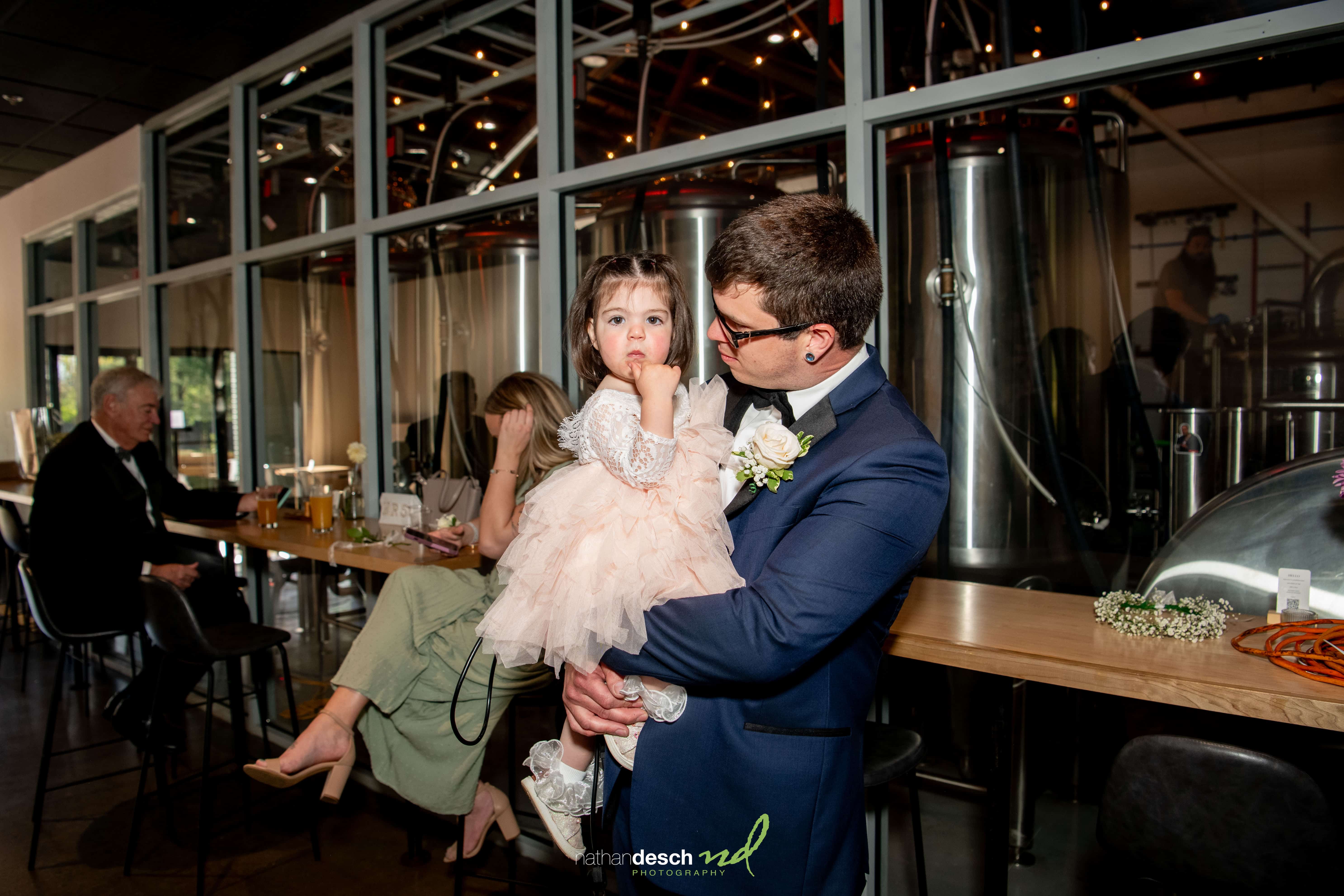 Groom in suit holding young girl in pink dress at brewery wedding venue with bar patrons and brewing tanks in background.