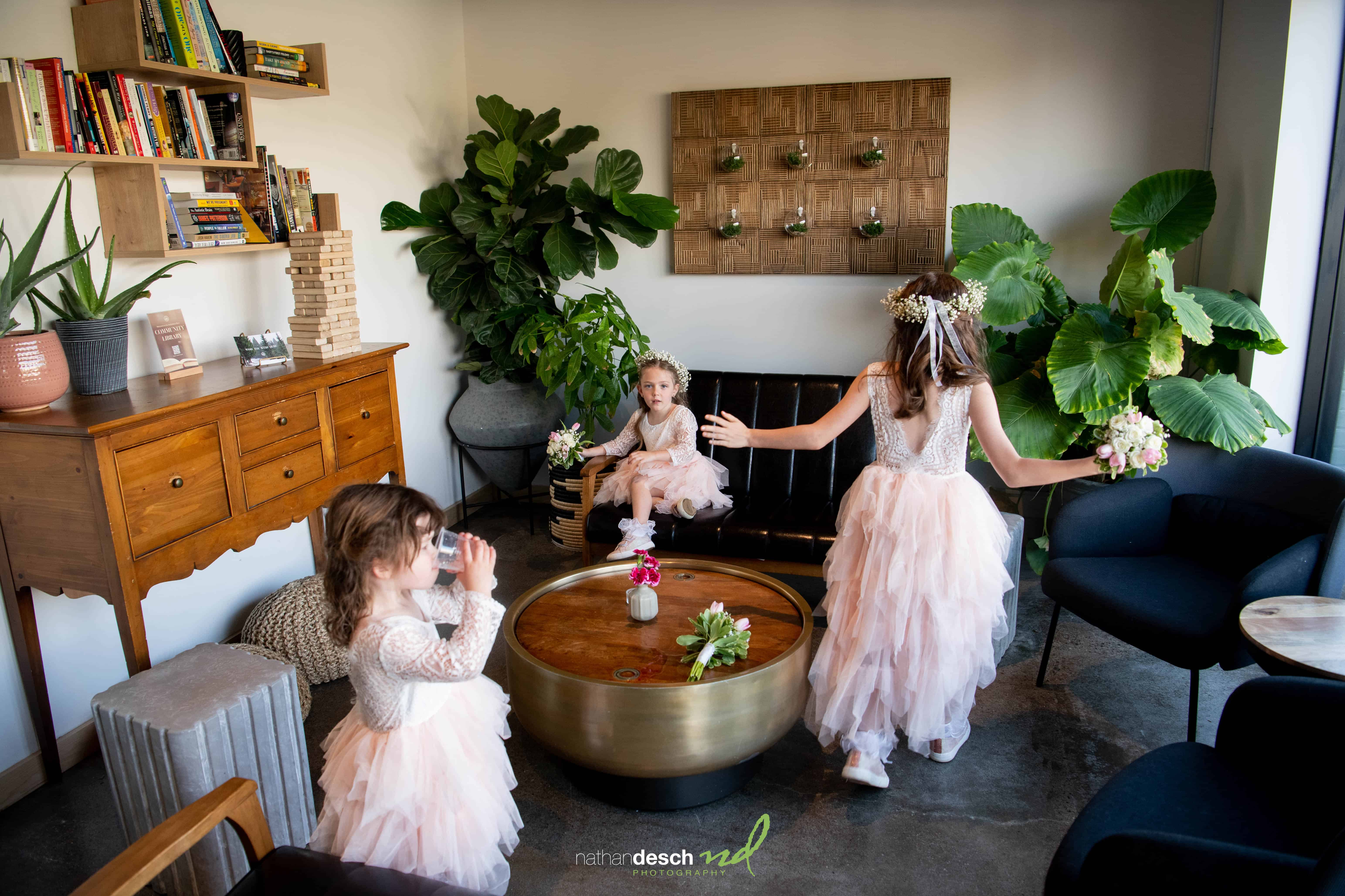 Three young girls in matching pink dresses play indoors with bouquets, surrounded by lush plants and elegant wedding decor.