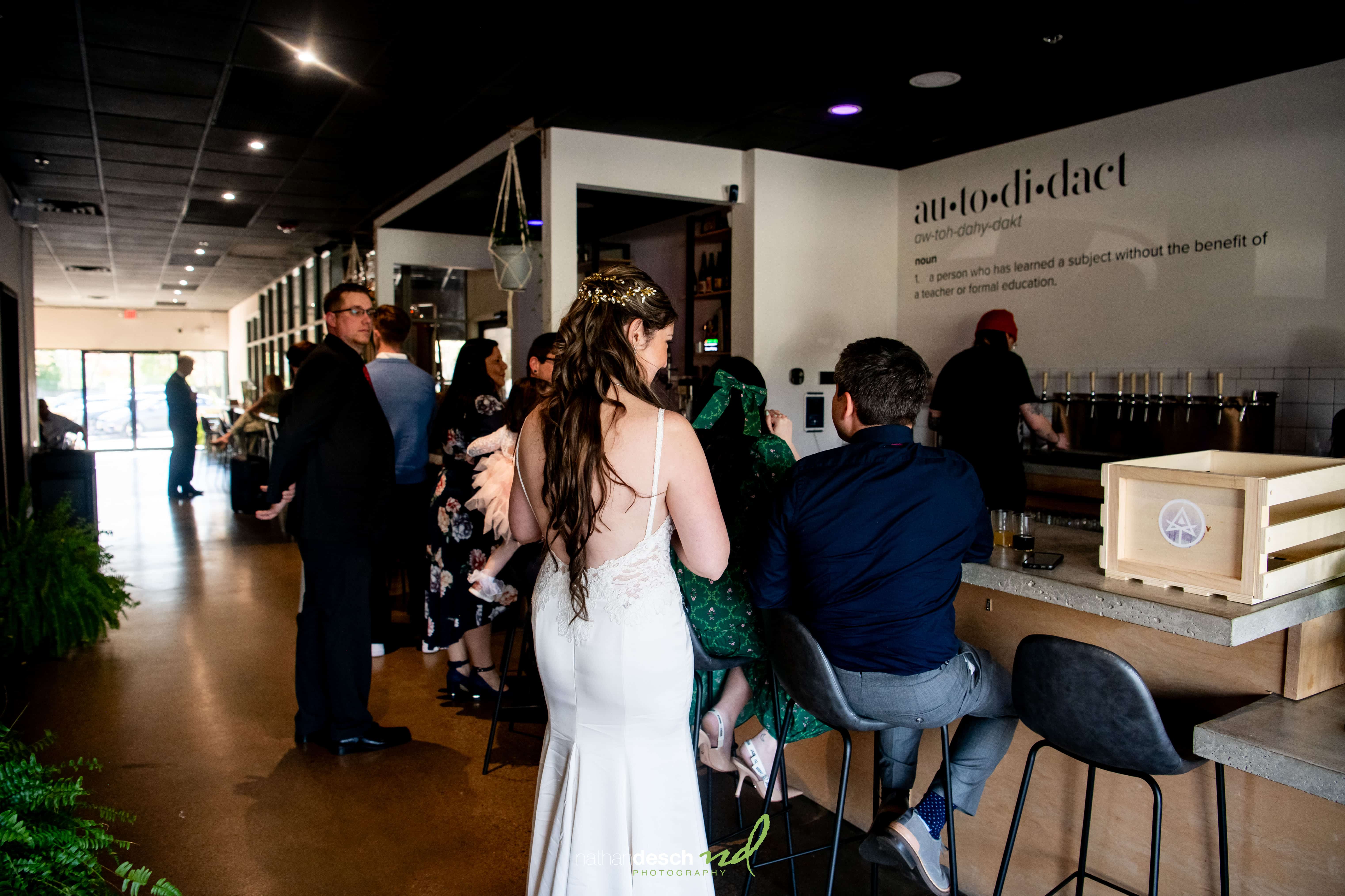 Bride Kaitlyn in white at modern, dimly lit wedding venue bar with guests, lush greenery, and wall sign in background.