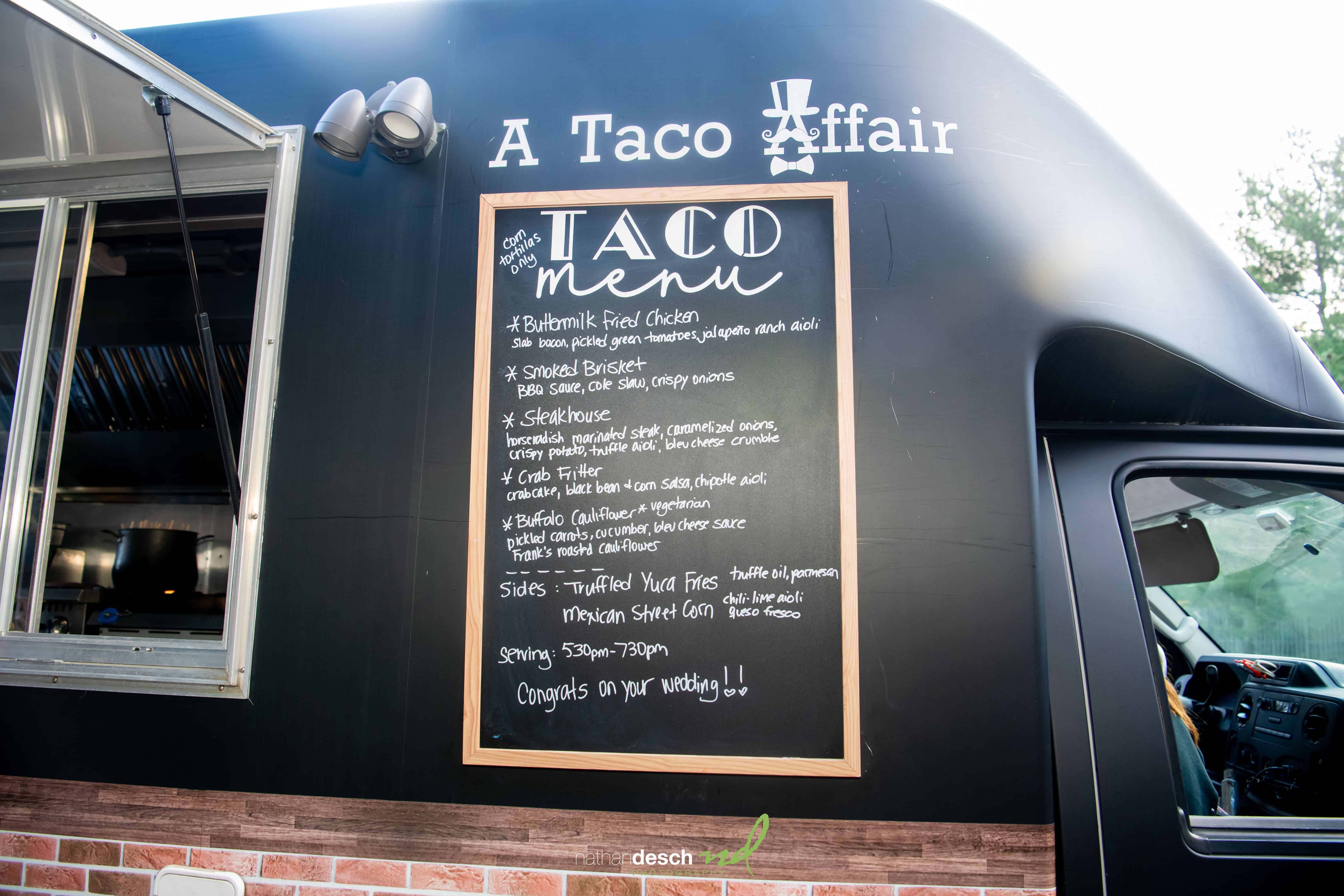 Black food truck labeled A Taco Affair with chalkboard menu and server Billy, surrounded by greenery at a wedding reception.