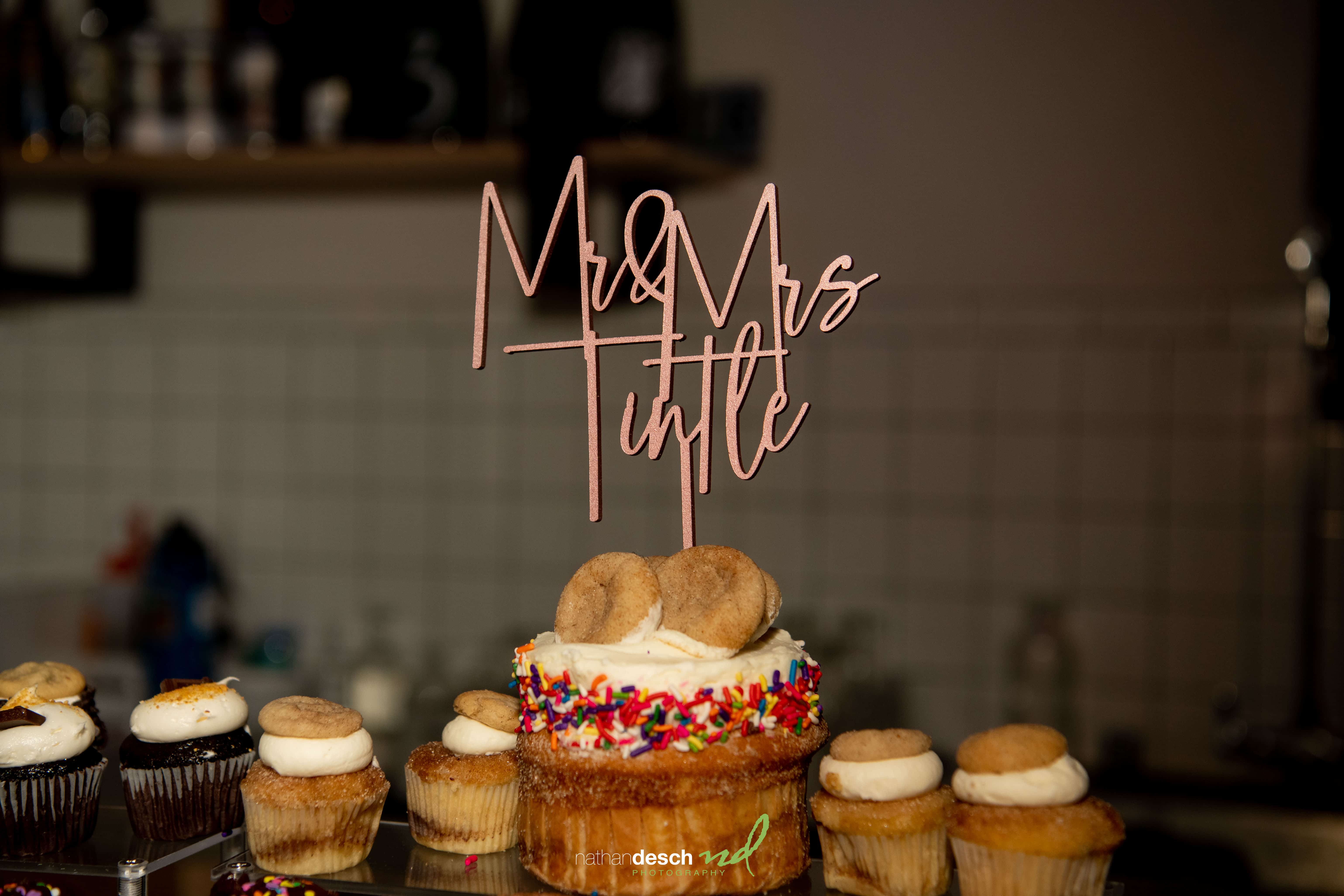 Cupcake with rainbow sprinkles and “Mr & Mrs Tuttle” topper, surrounded by cookies and cupcakes for Billy and Kaitlyns wedding.