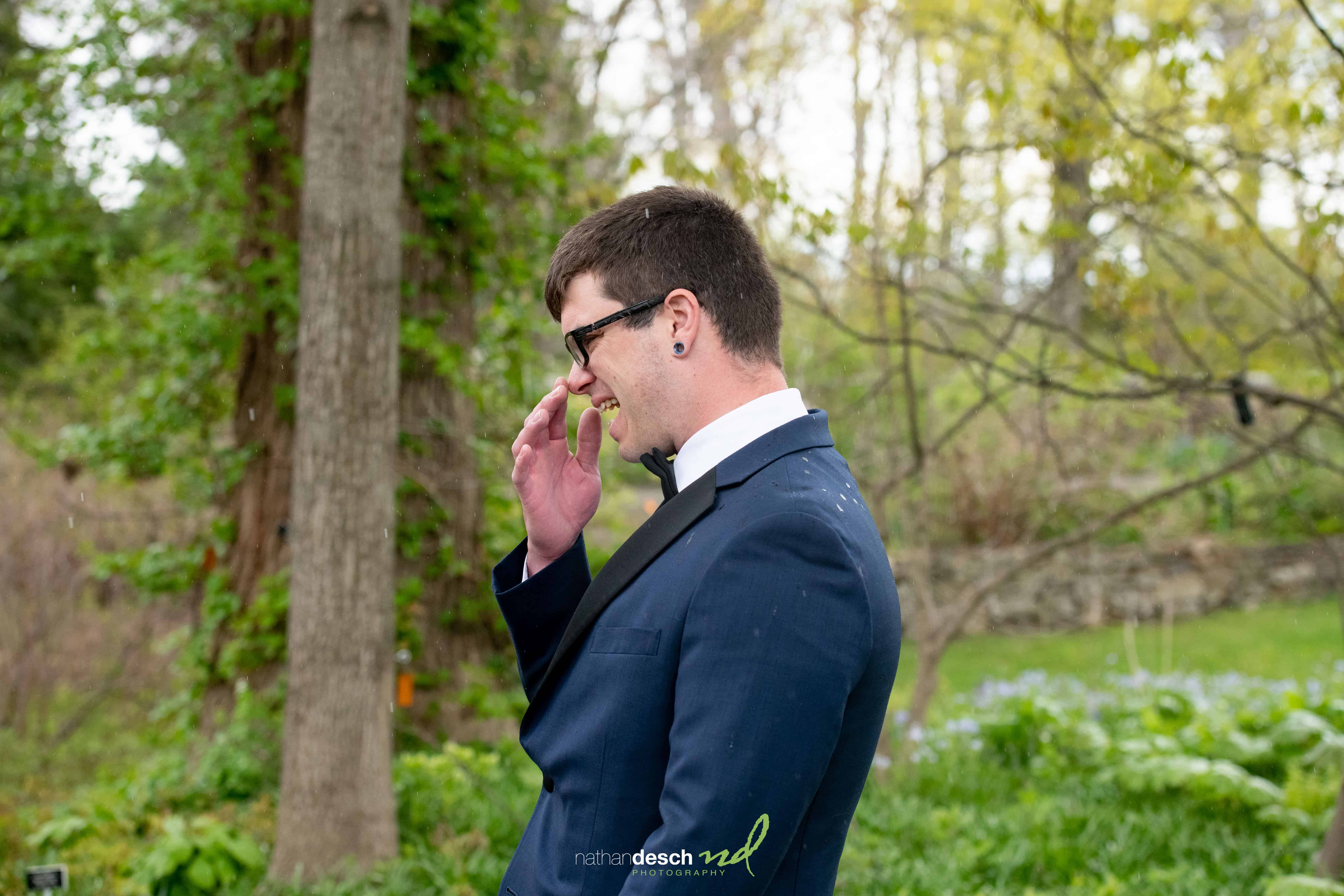 Groom in navy suit smiles outdoors at Frelinghuysen Arboretum wedding, surrounded by lush greenery and trees.