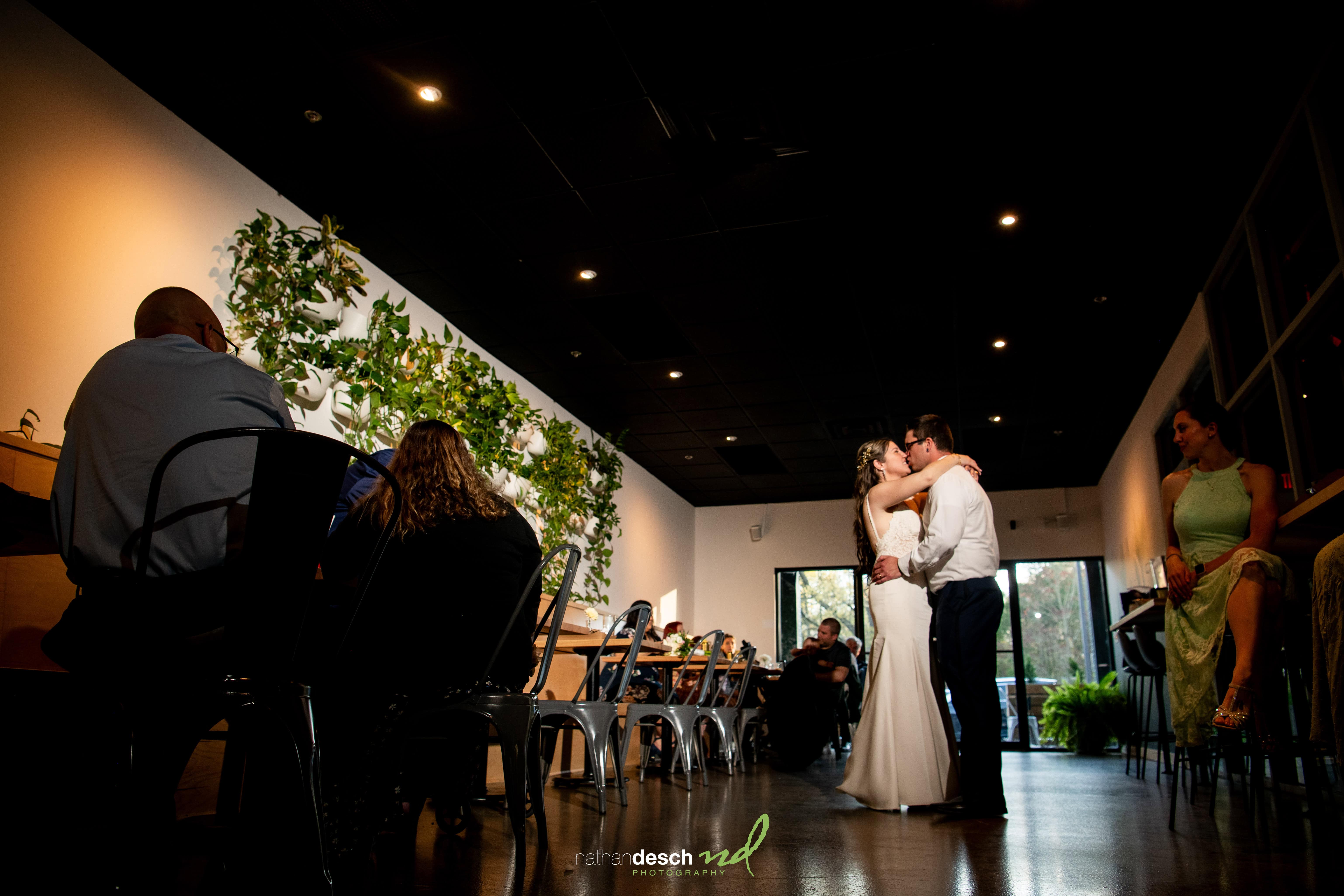 Bride and groom share a kiss on the dance floor in a modern, sunlit wedding venue with greenery decor and watching guests.
