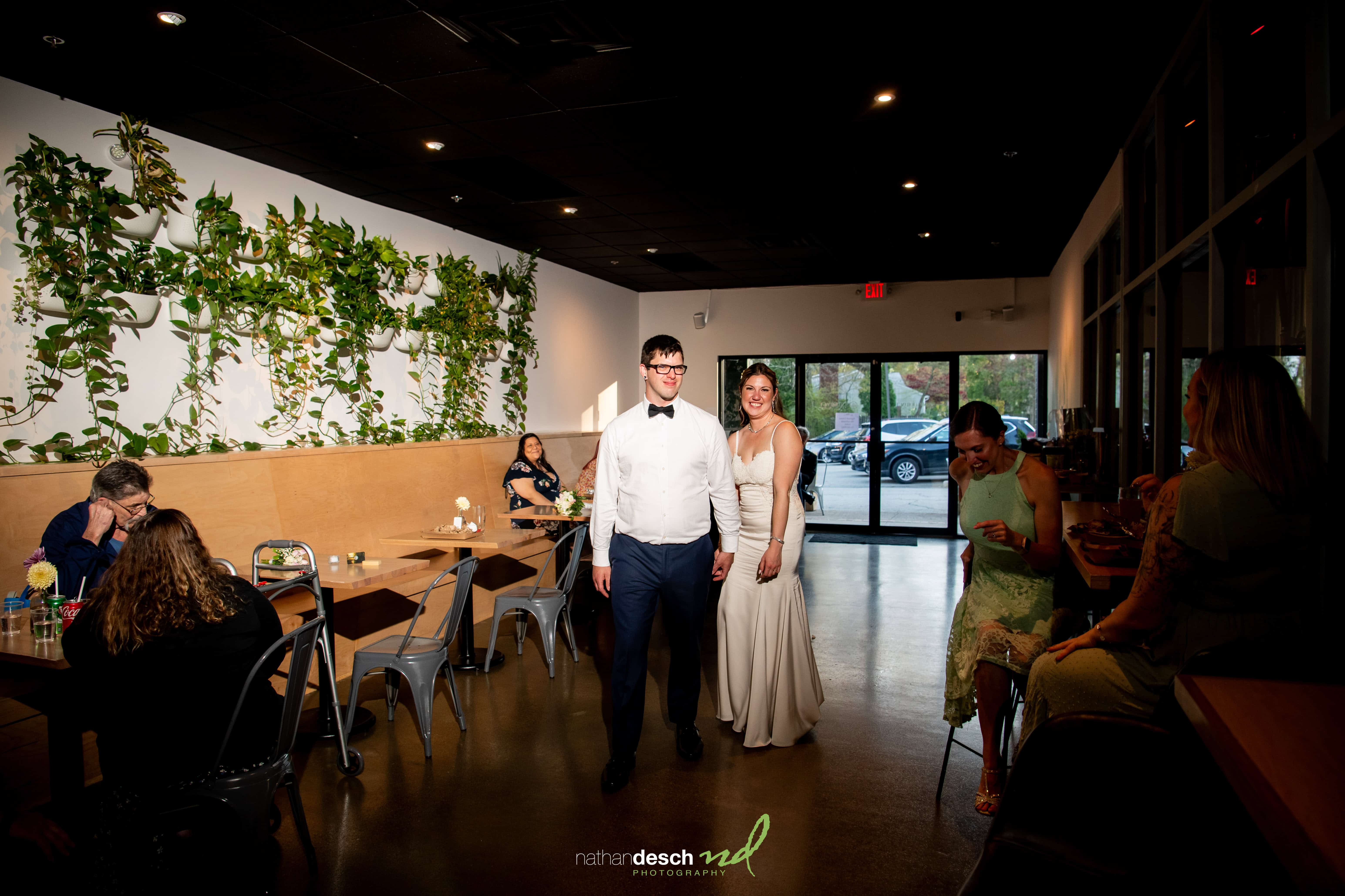 Bride and groom smiling, holding hands, enter a modern plant-filled restaurant as guests watch and take photos, warm lighting.