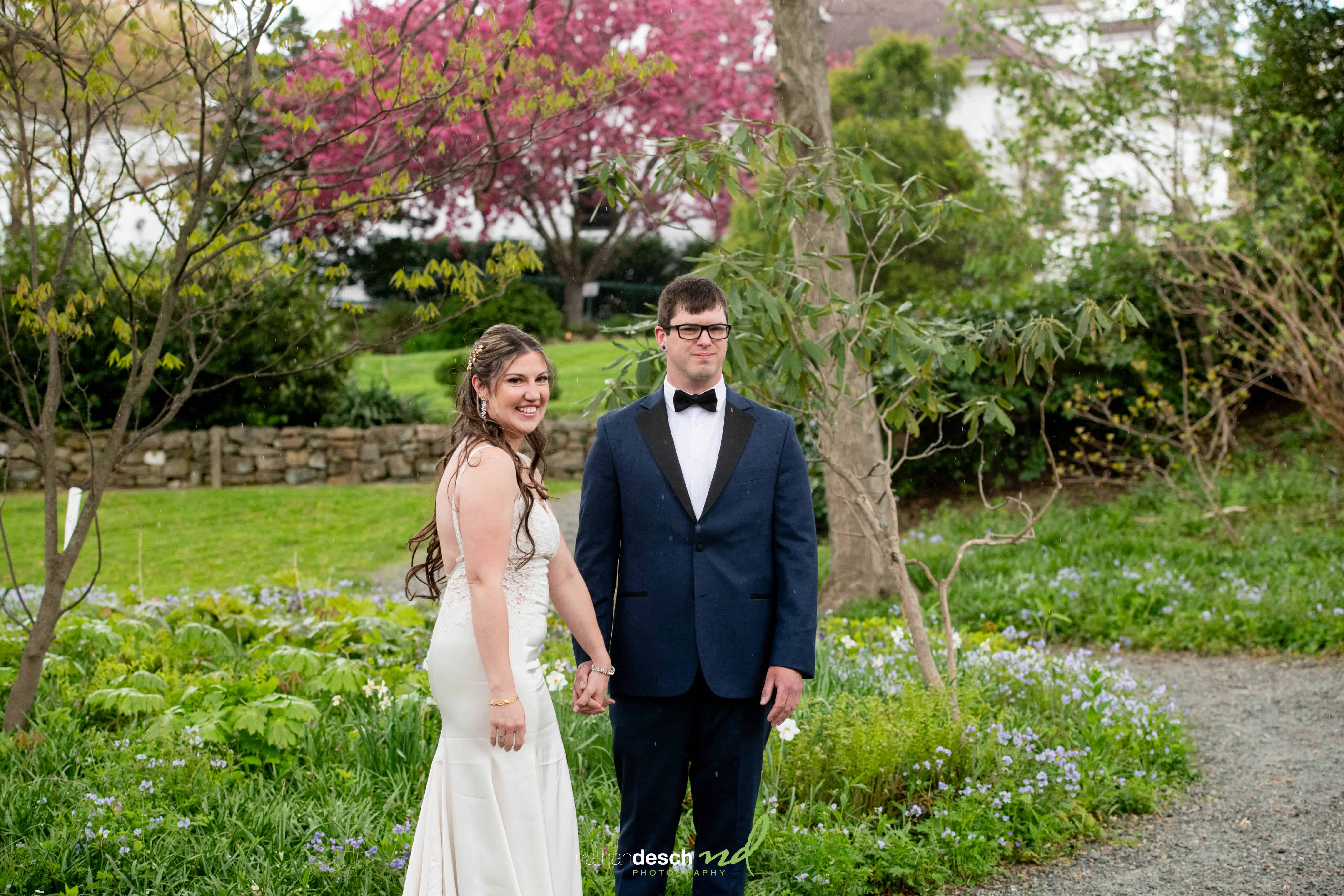 Bride in white dress and groom in navy suit hold hands, smiling in a lush garden at Frelinghuysen Arboretum wedding.
