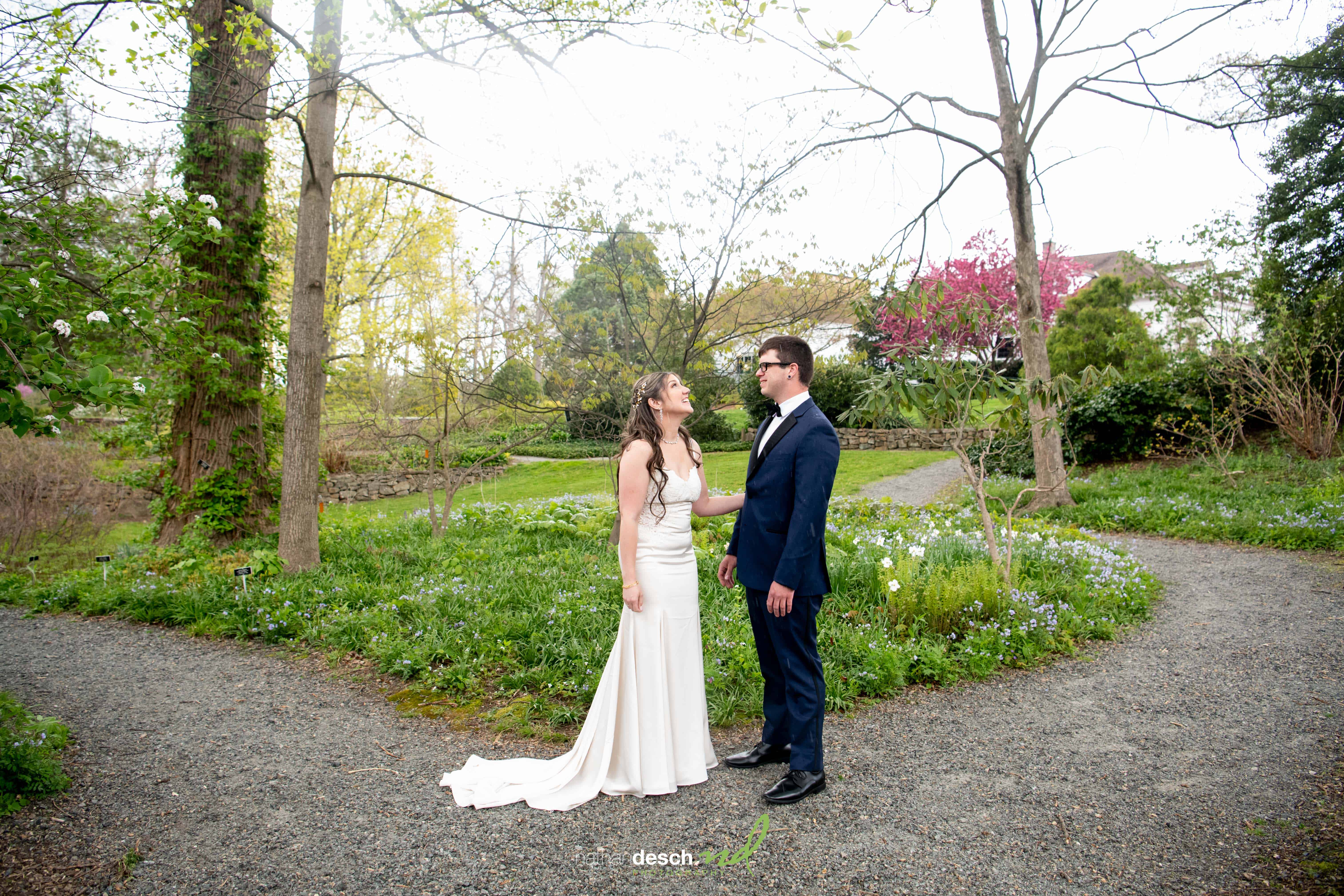 Bride and groom stand on a gravel path at Frelinghuysen Arboretum, surrounded by lush greenery and blooming flowers.